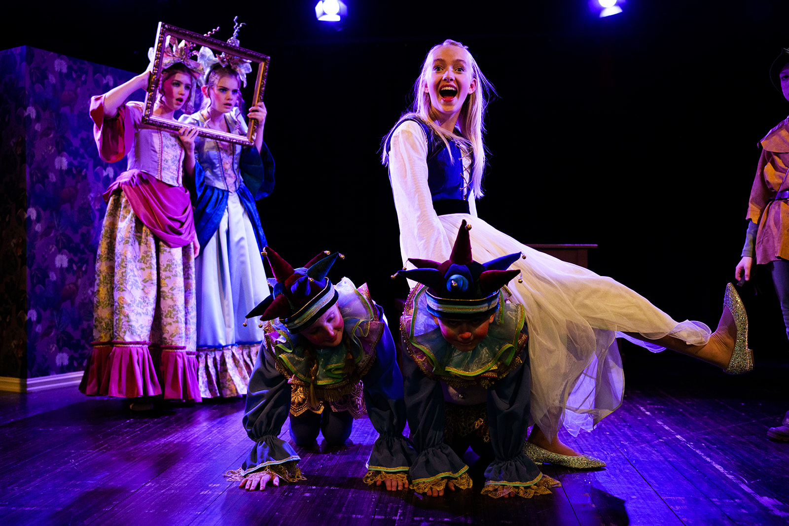 Jente som sitter på ryggen til en hoffnarr og to jenter ikledd prinsessekostymer som ser gjennom en bilderamme med forbausede blikk