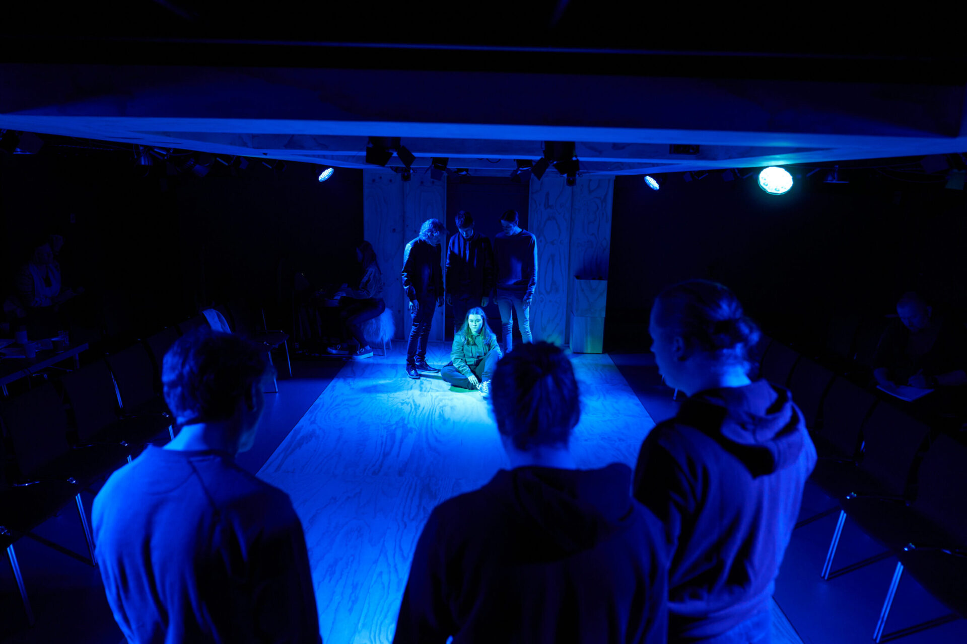 Ei jente sitter på gulvet. Tre gutter står bak. Tre gutter står foran og ser på.