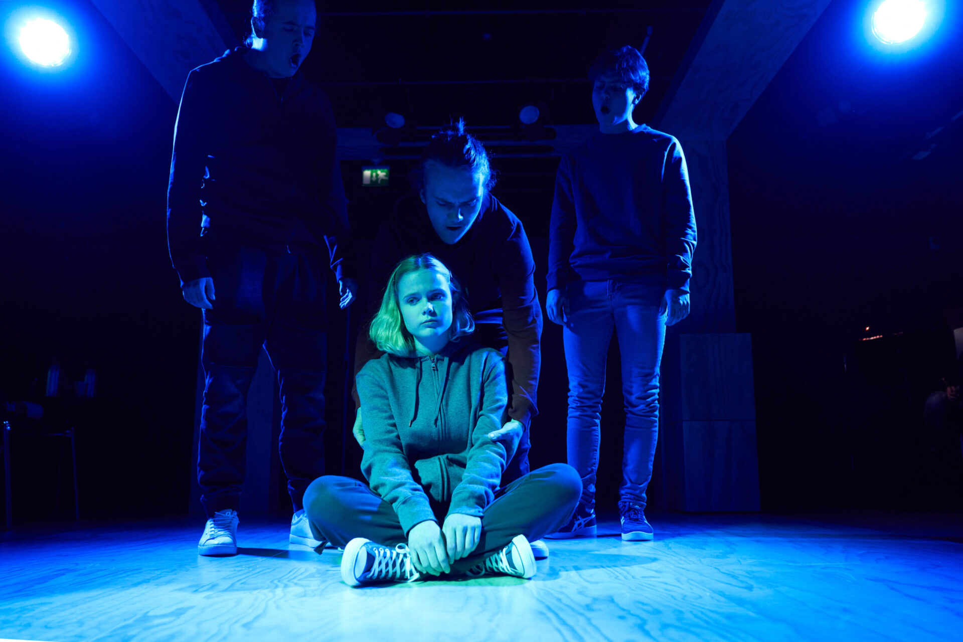 Ei jente sitter på gulvet mens tre gutter står bak.