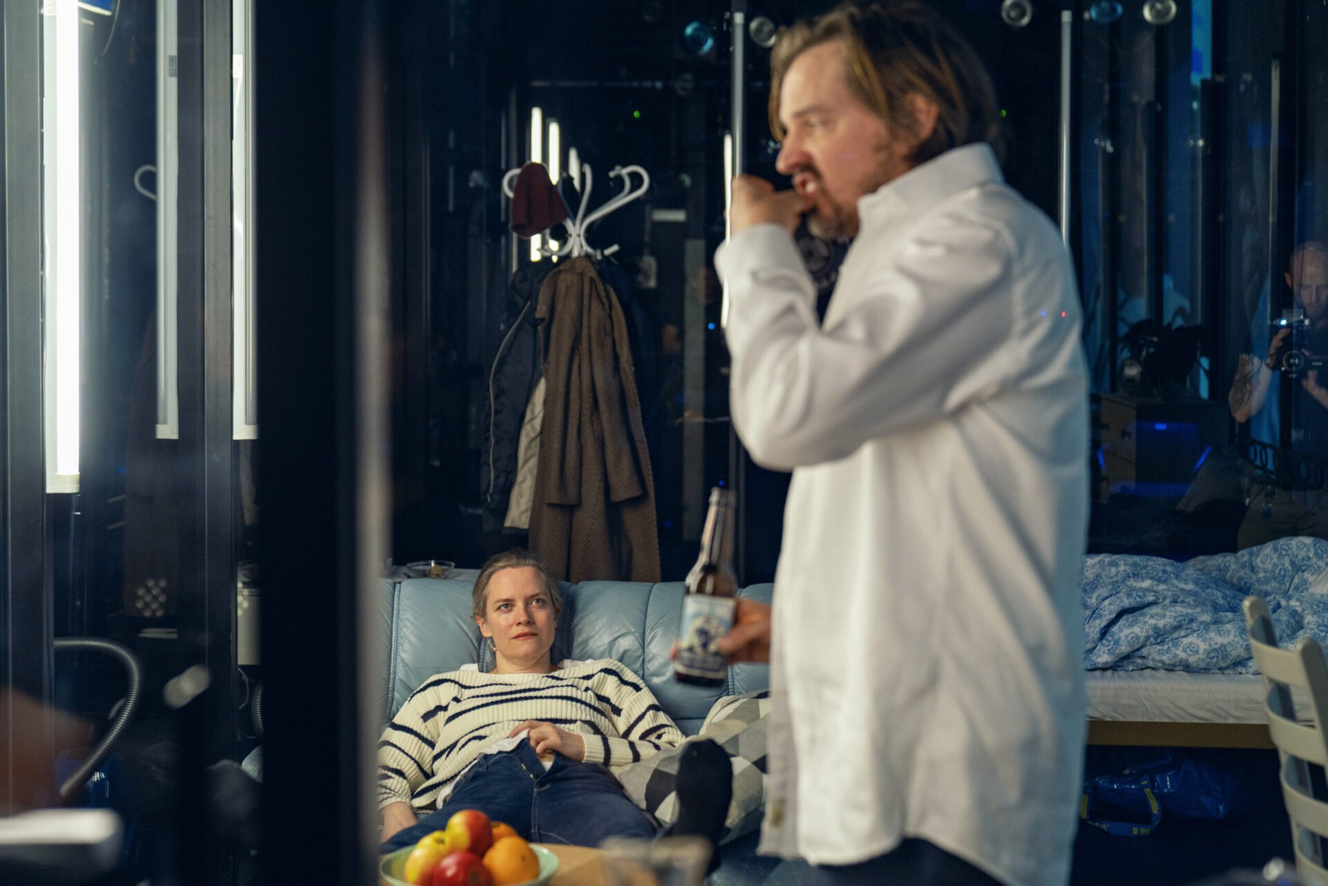 En kvinne ligger i en sofa mens en mann i hvit skjorte spiser en brødskive.