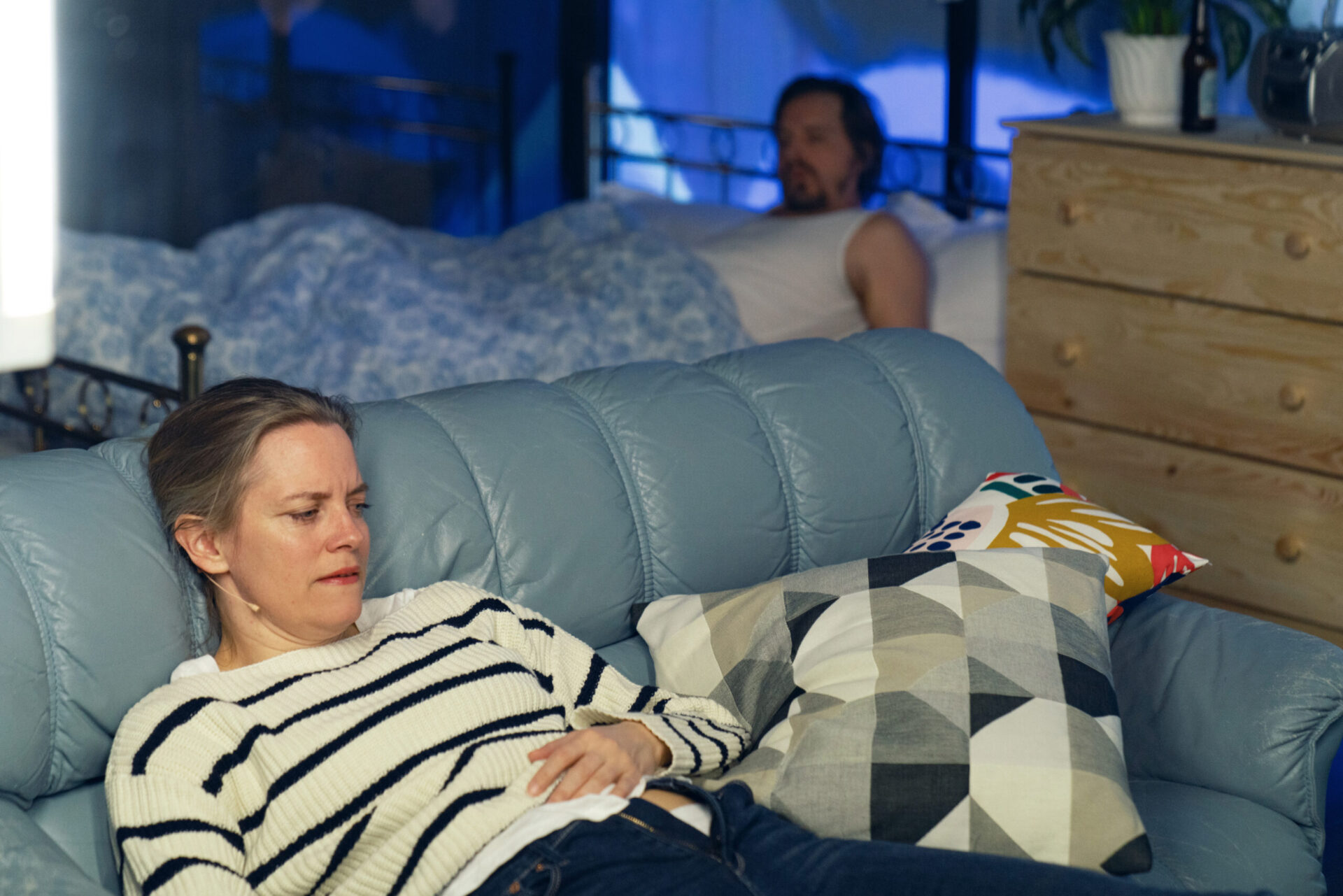 En kvinne ligger på en sofa, i bakgrunnen en mann i en seng.