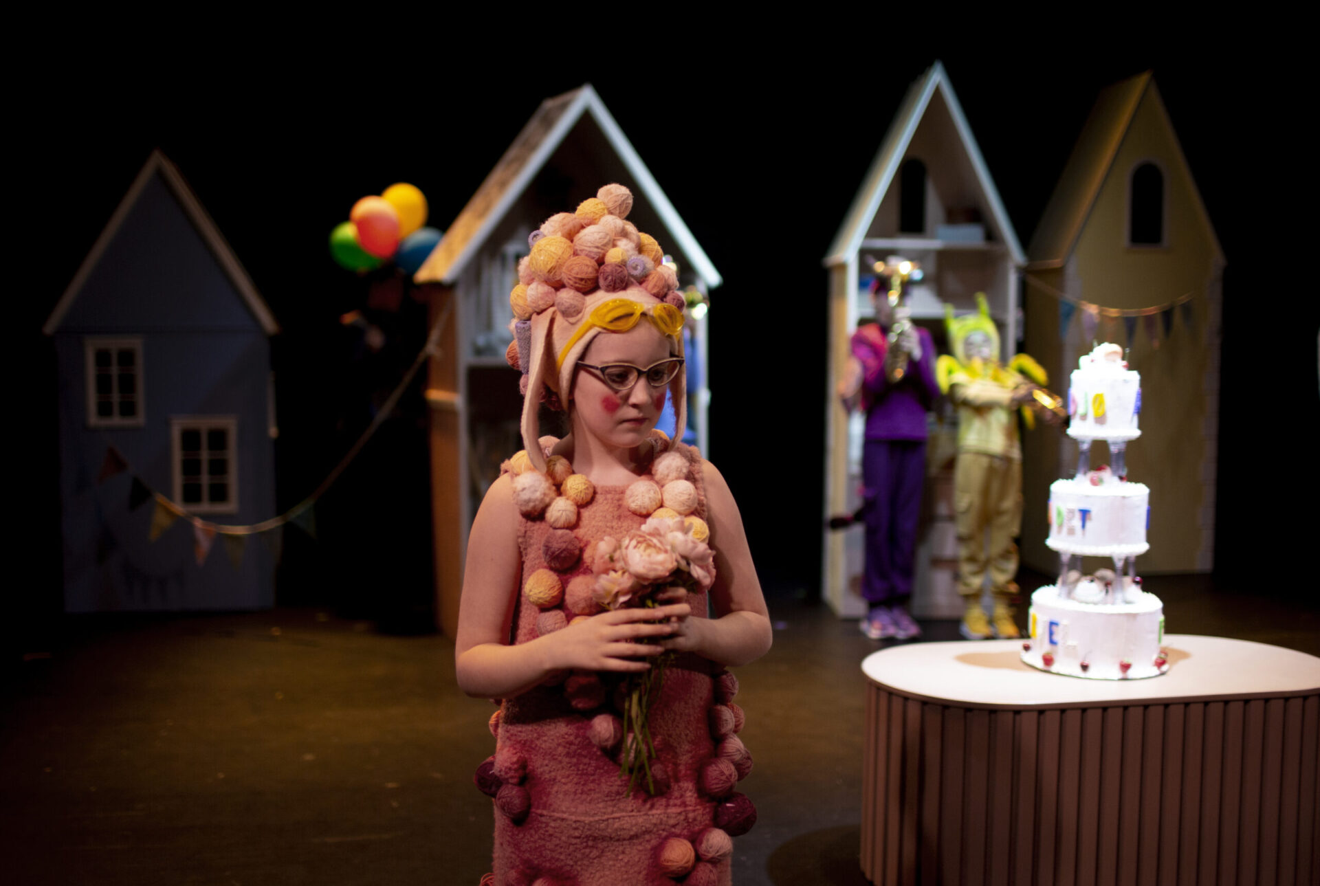 En jente i rosa kostyme med masse garnnøster står med blomster foran en stor kake.