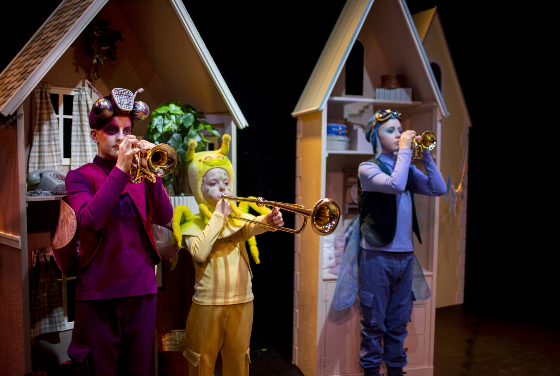 Tre unge skuespillere i fargerike kostymer spiller på blåseinstrumenter.