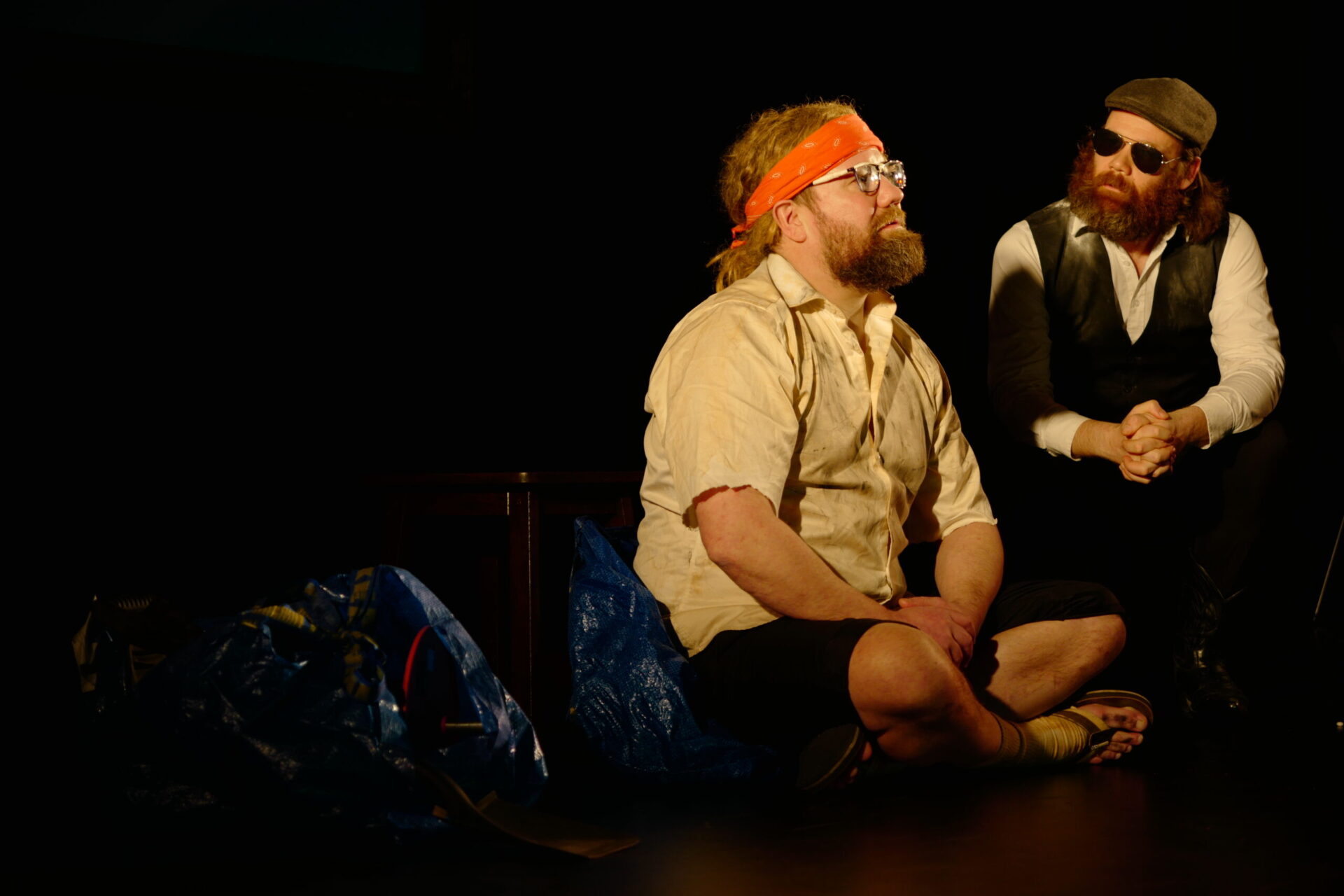 En mann sitter i yogastilling med orange pannebånd på seg. En annen mann med solbriller sitter på huk ved siden av.