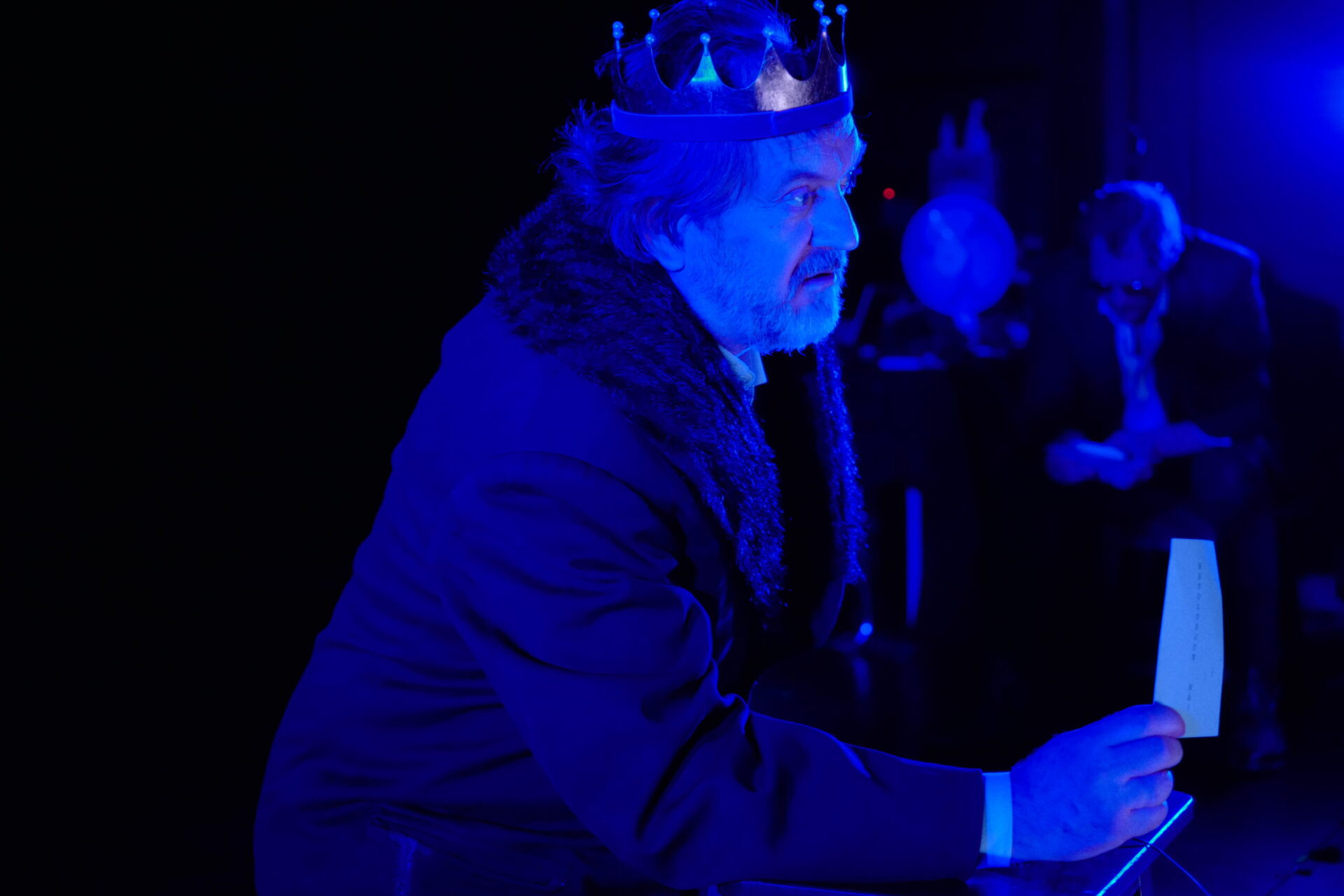 En mann med kongekrone holder et papirark mens en annen mann sitter i bakgrunnen.