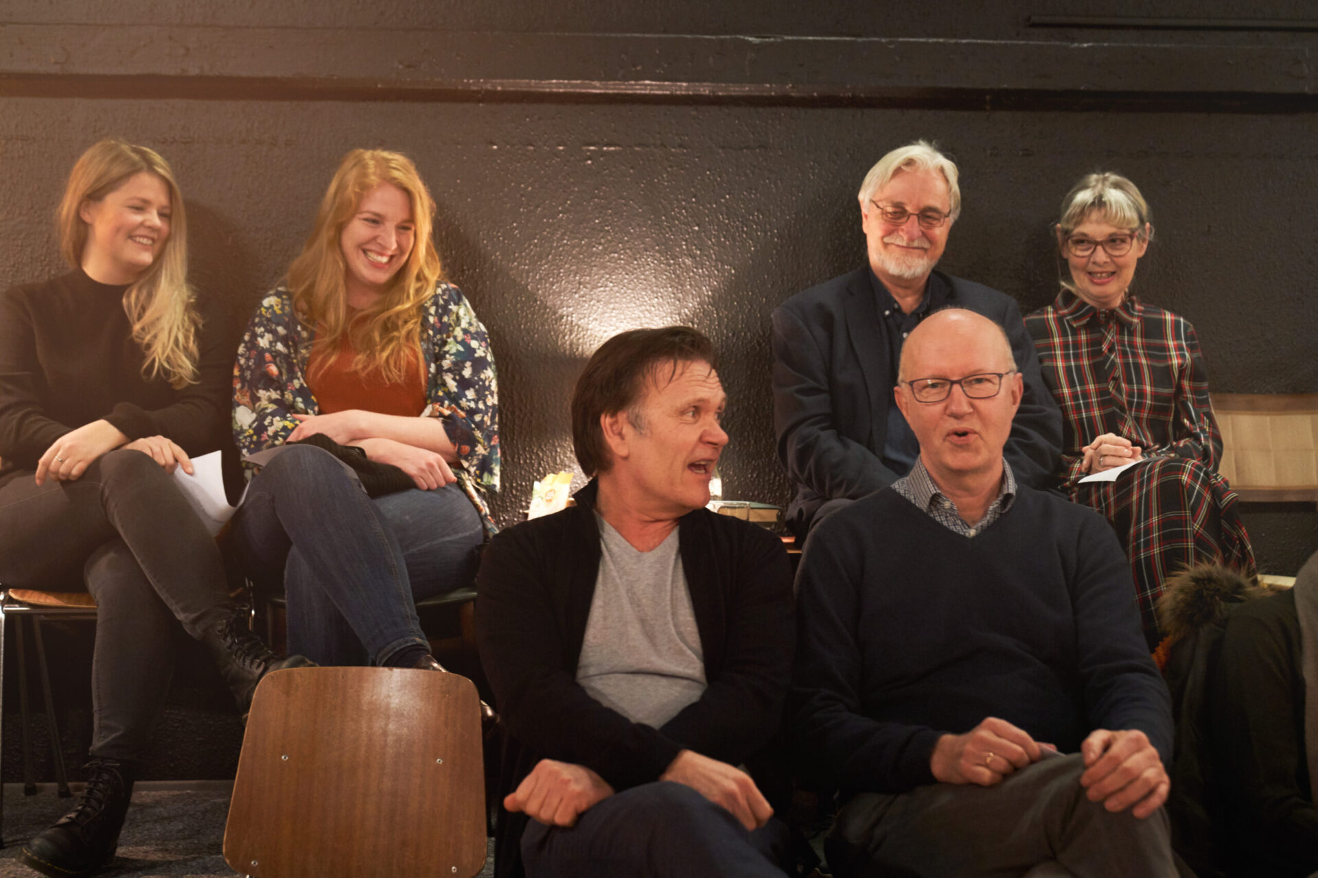 Espen Hana sitter ved siden av en eldre mann og snakker men noen andre sitter bak og ler.