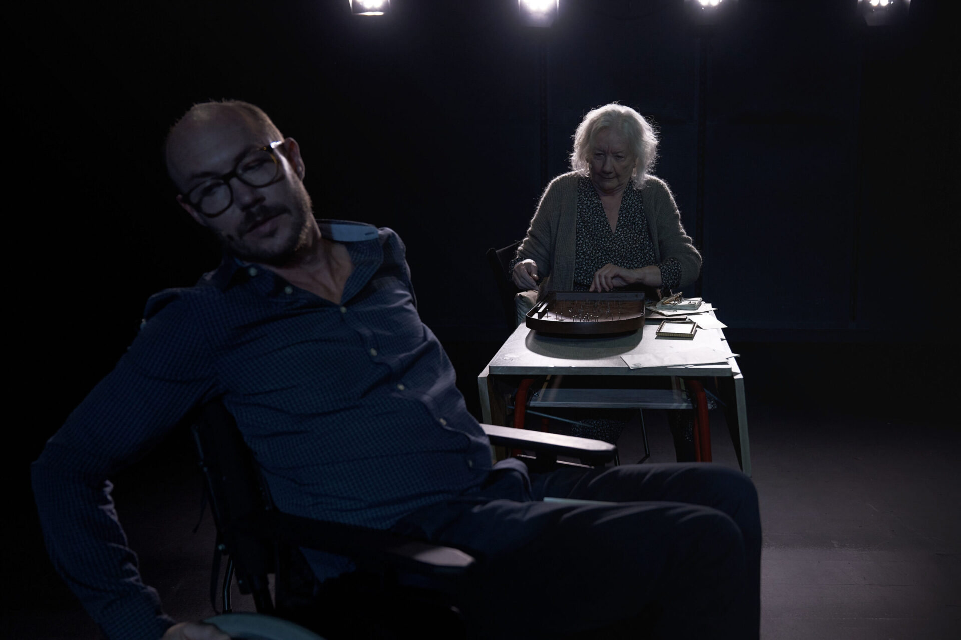 En eldre kvinne sitter med et spill, mens en mann i rullestol ønsker å komme seg vekk