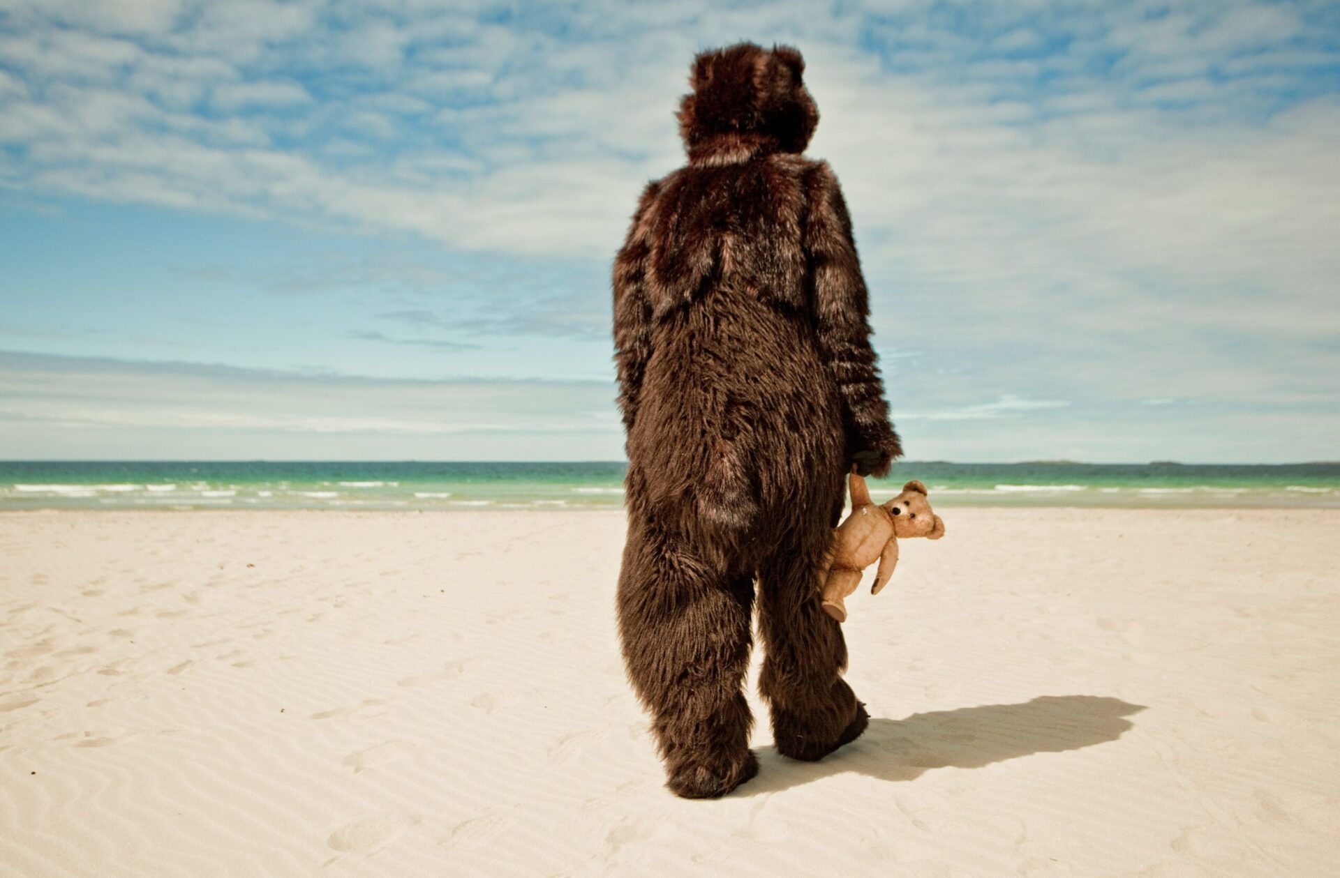En bjørn med en teddybjørn i hånden på en strand.
