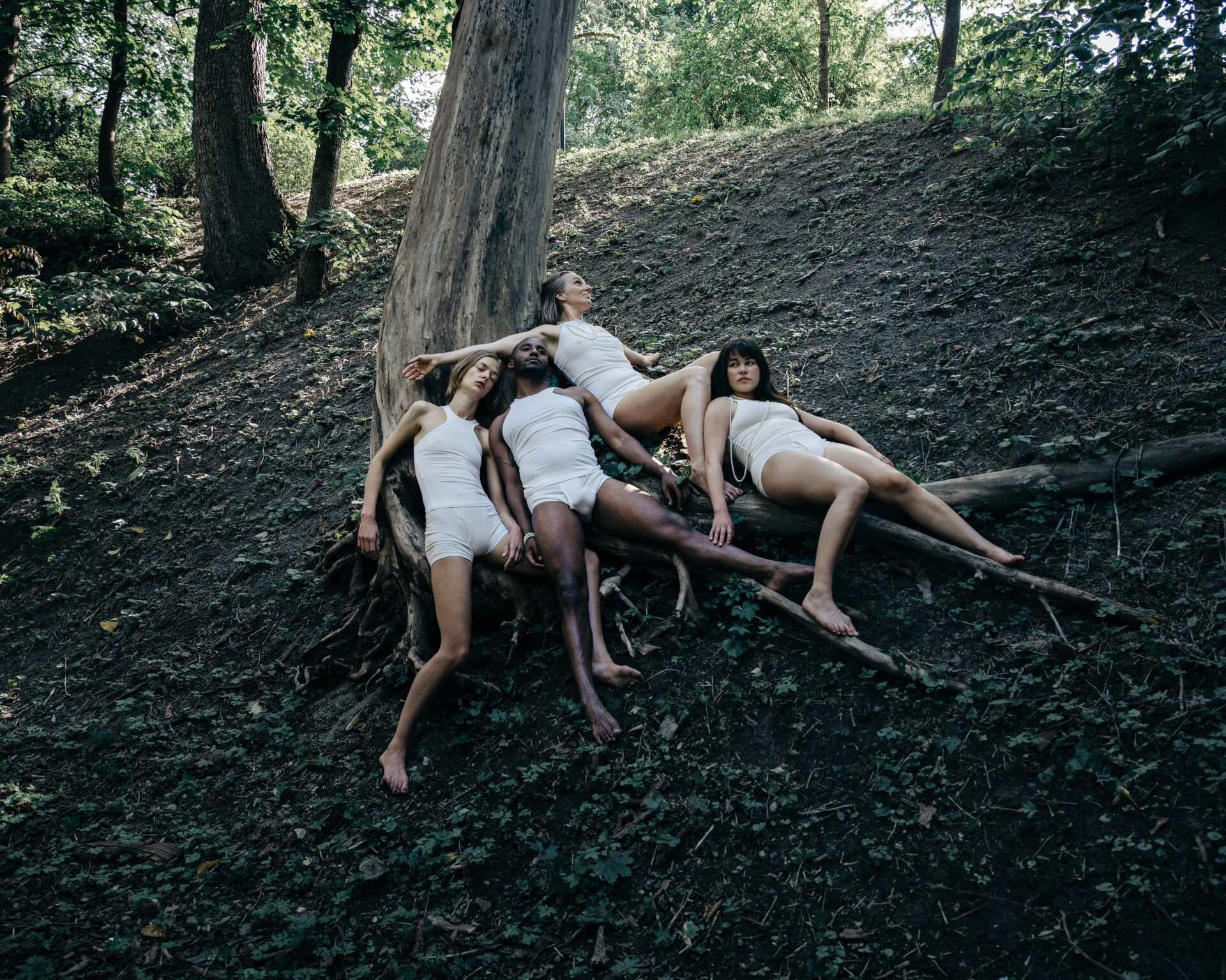 Mennesker i hvite klær som ligger strødd på skogbunnen under et tre.