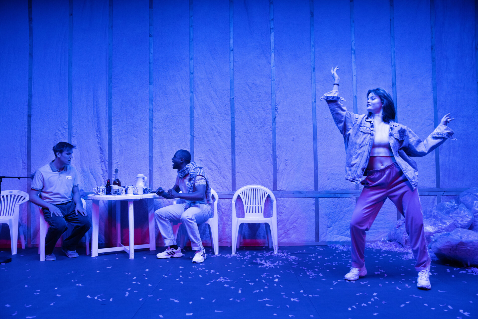 En ung kvinne i magetopp danser mens to menn sitter ved et bord og drikker.