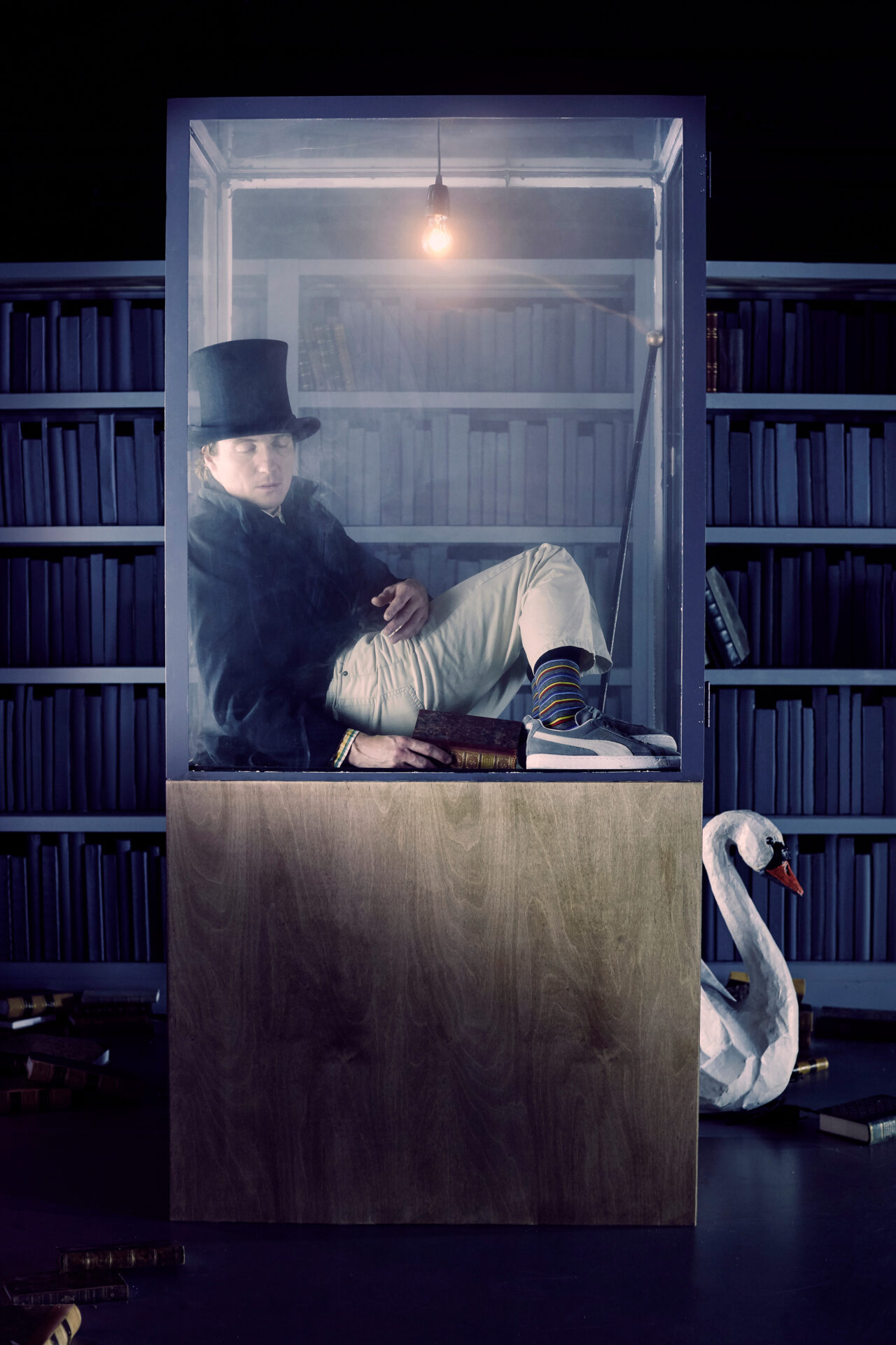 en mann i et blått bibliotek, inne i en glassmonter, med en flosshatt på hode, og en svane på gulvet