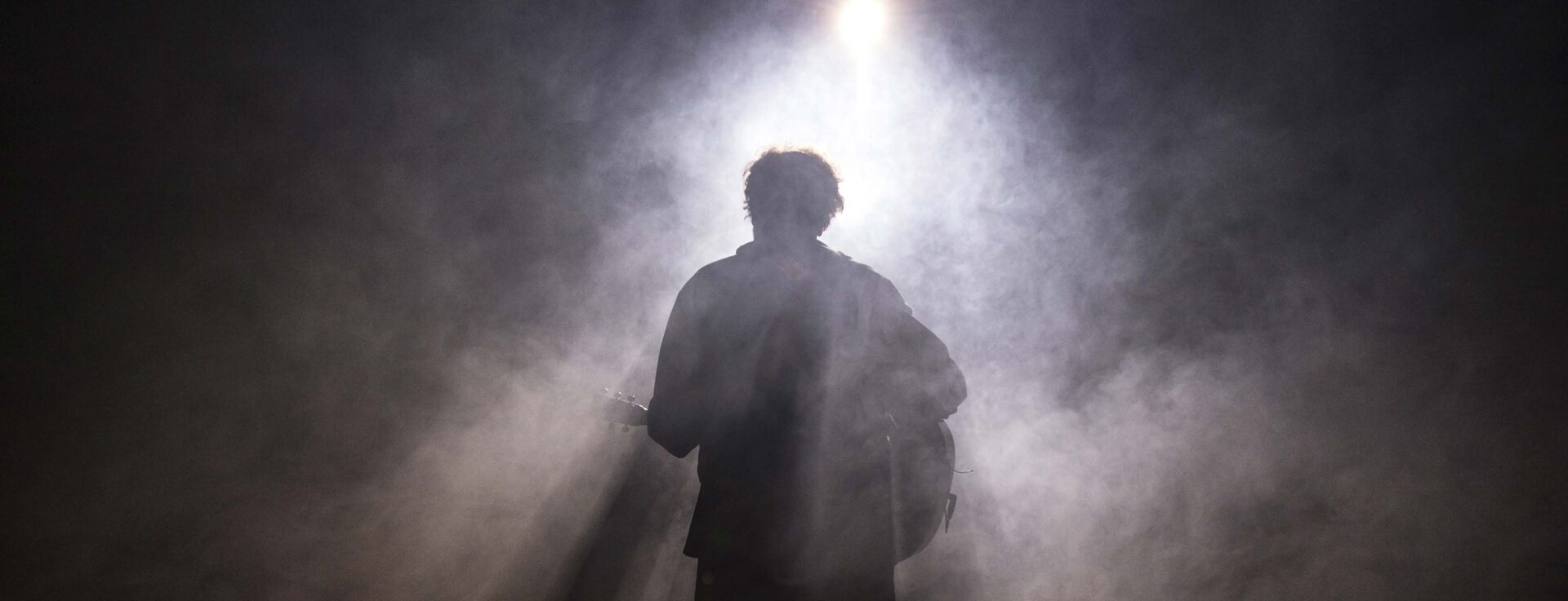 Ledig stilling hos Rogaland Teater som lystekniker illustrert med bilde av mann med gitar sett bakfra på en scene, i en spotlight.