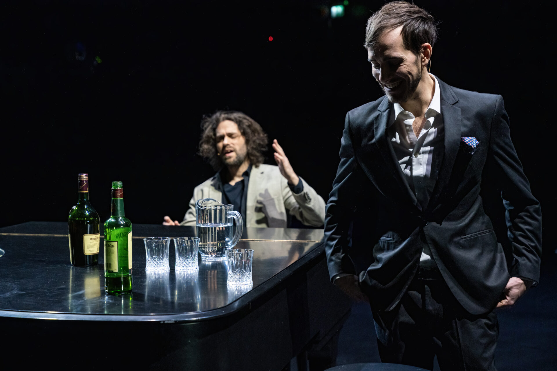 Et piano med wiskyflasker. En mann som sitter full ved pianoet og en som står ved siden av og ler.