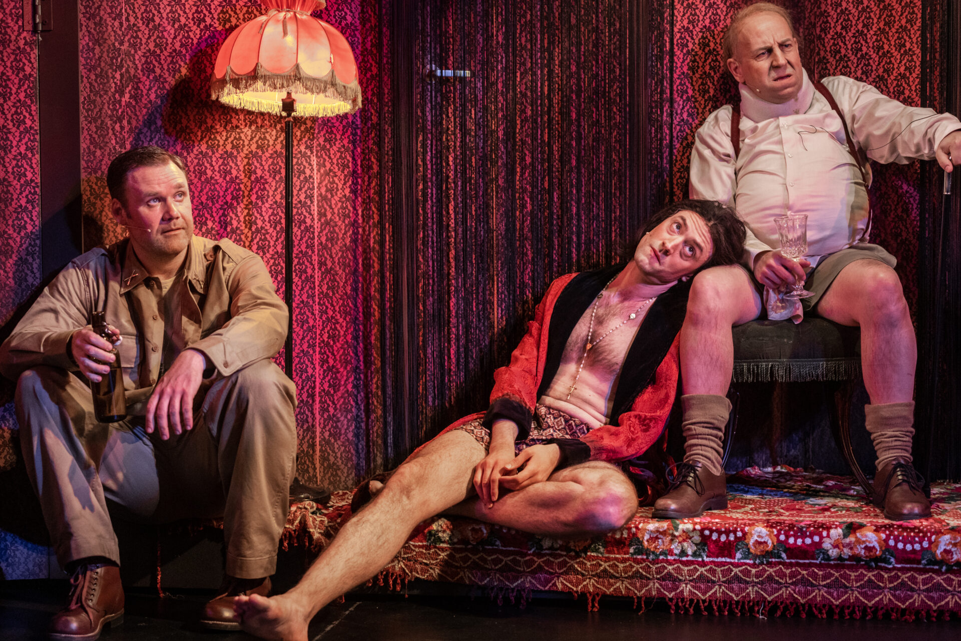 En mann i militærtøy, en mann i kvinnetøy og en mann med nakkekrage sitter i et bordell.