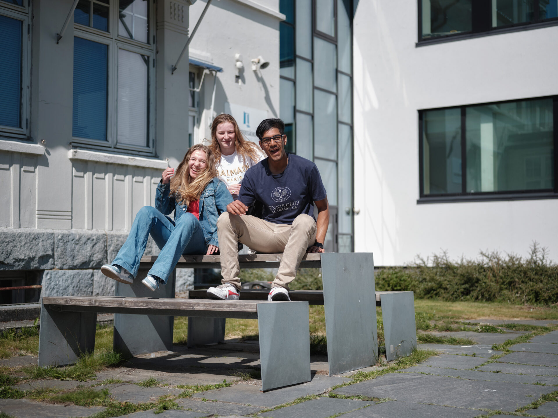 Tre smilende ungdommer som sitter på en benk utenfor skolen