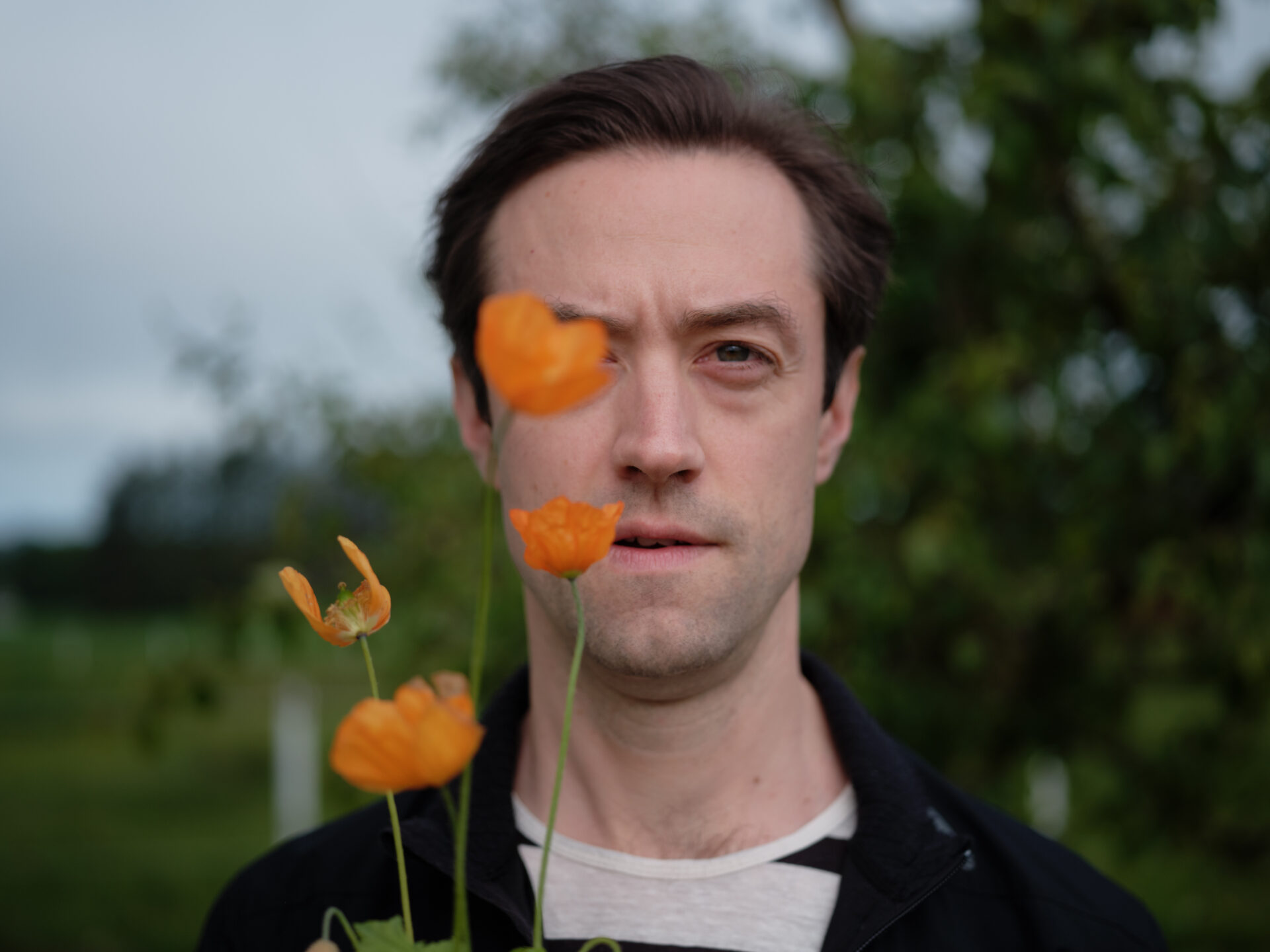 Mann ser inn i kamera, holder blomst foran det ene øyet