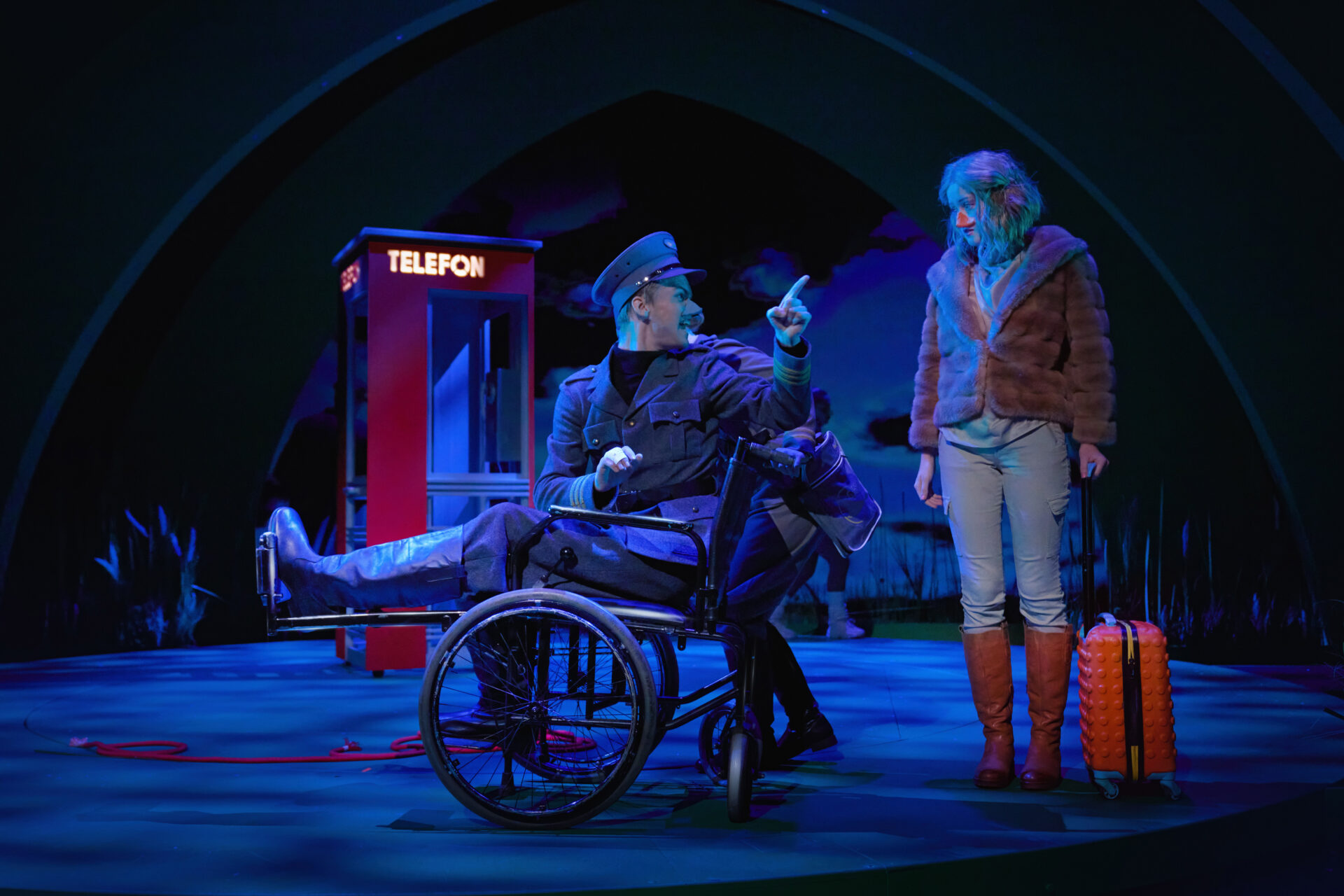 et barn sitter i rullestol, peker på annet barn som står med koffert