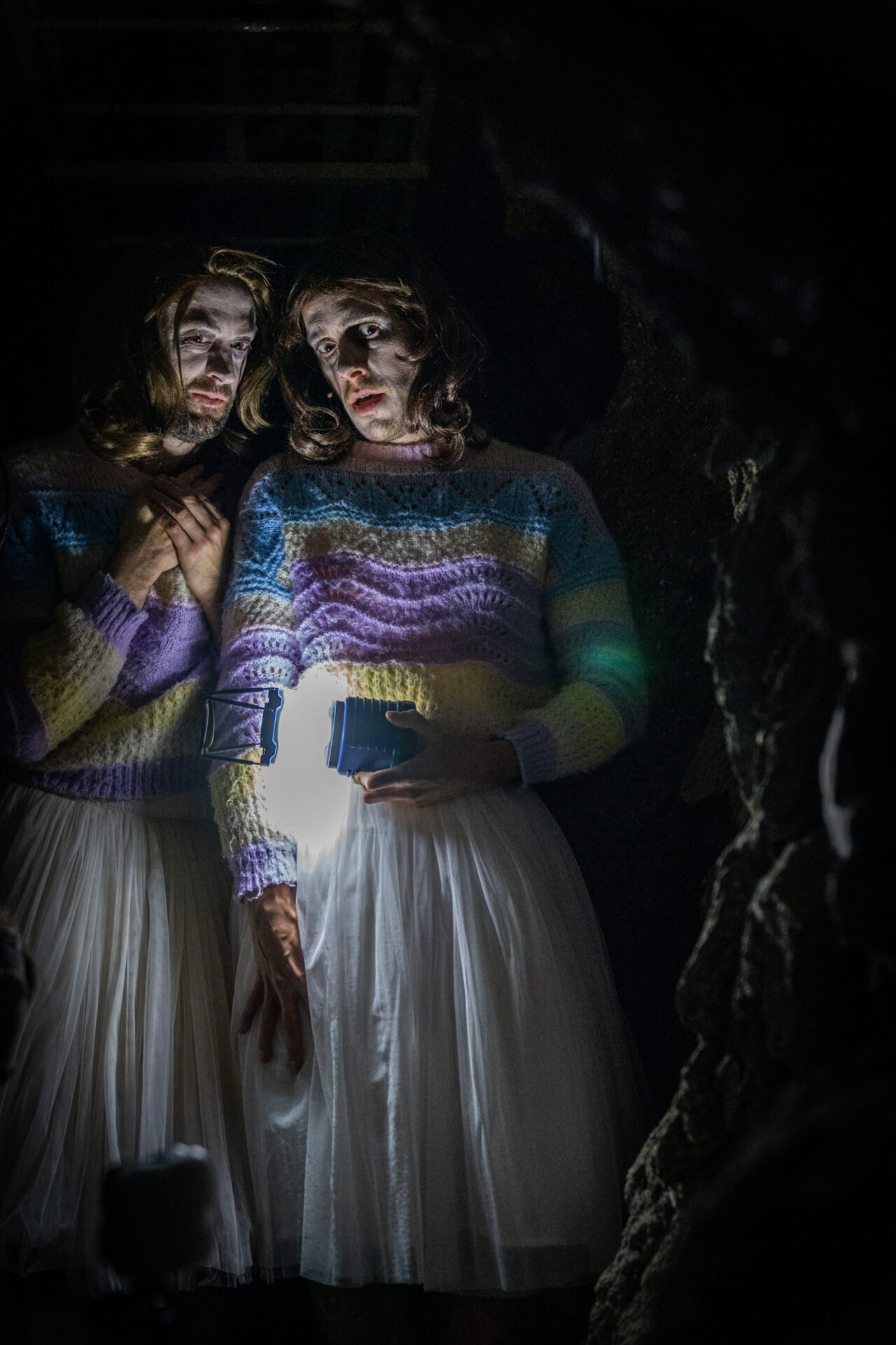 To menn med parykk kledd ut som jenter, i en mørk grotte