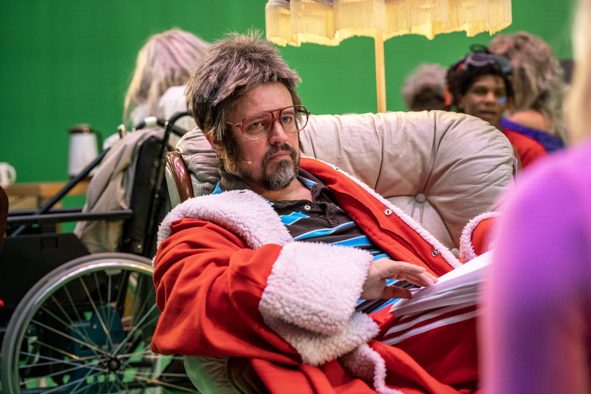 En mann med skjegg, briller, grilldress og julenissefrakk, som sitter bakoverlent i en gammel skinnstol