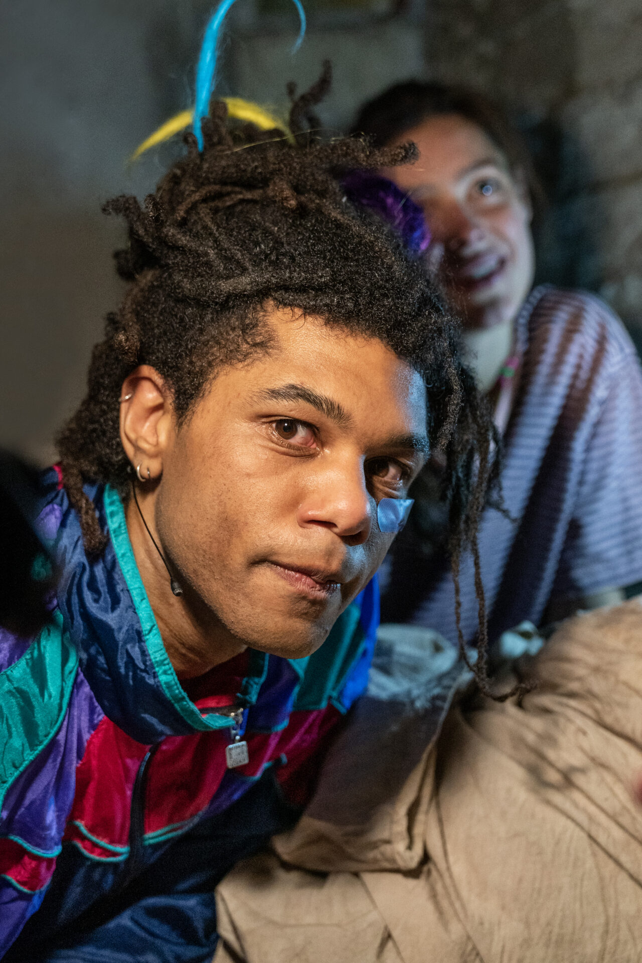 en mann som titter rett på kameraet, og sitter i en kjeller med en kvinne i bakgrunnen