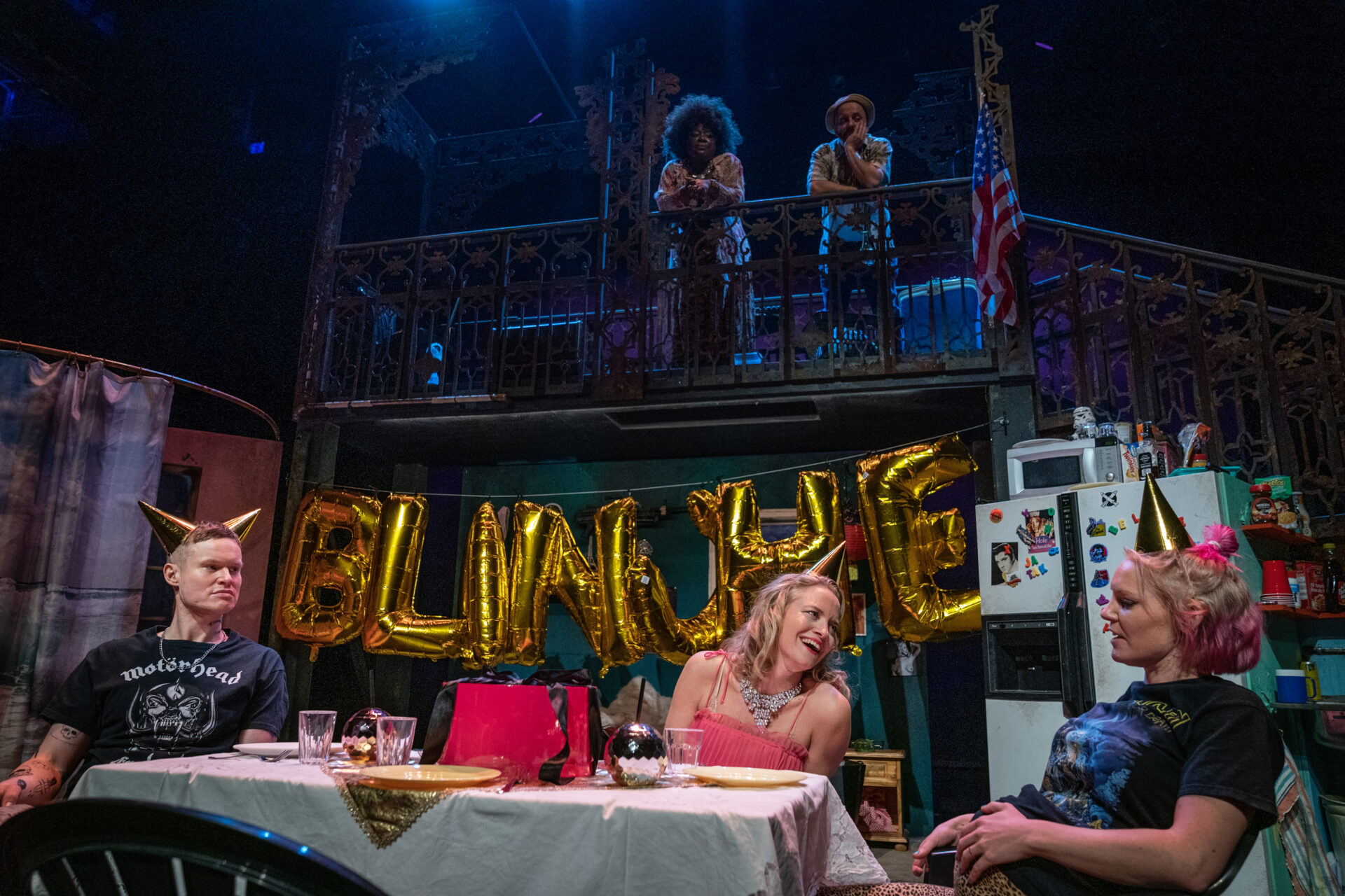 I et bursdagsselskap sitter en mann og to kvinner med papairhatt på hodet ved et bord og i bakgrunnen på en trappesats ser en mann og en kvinne ned de tre.