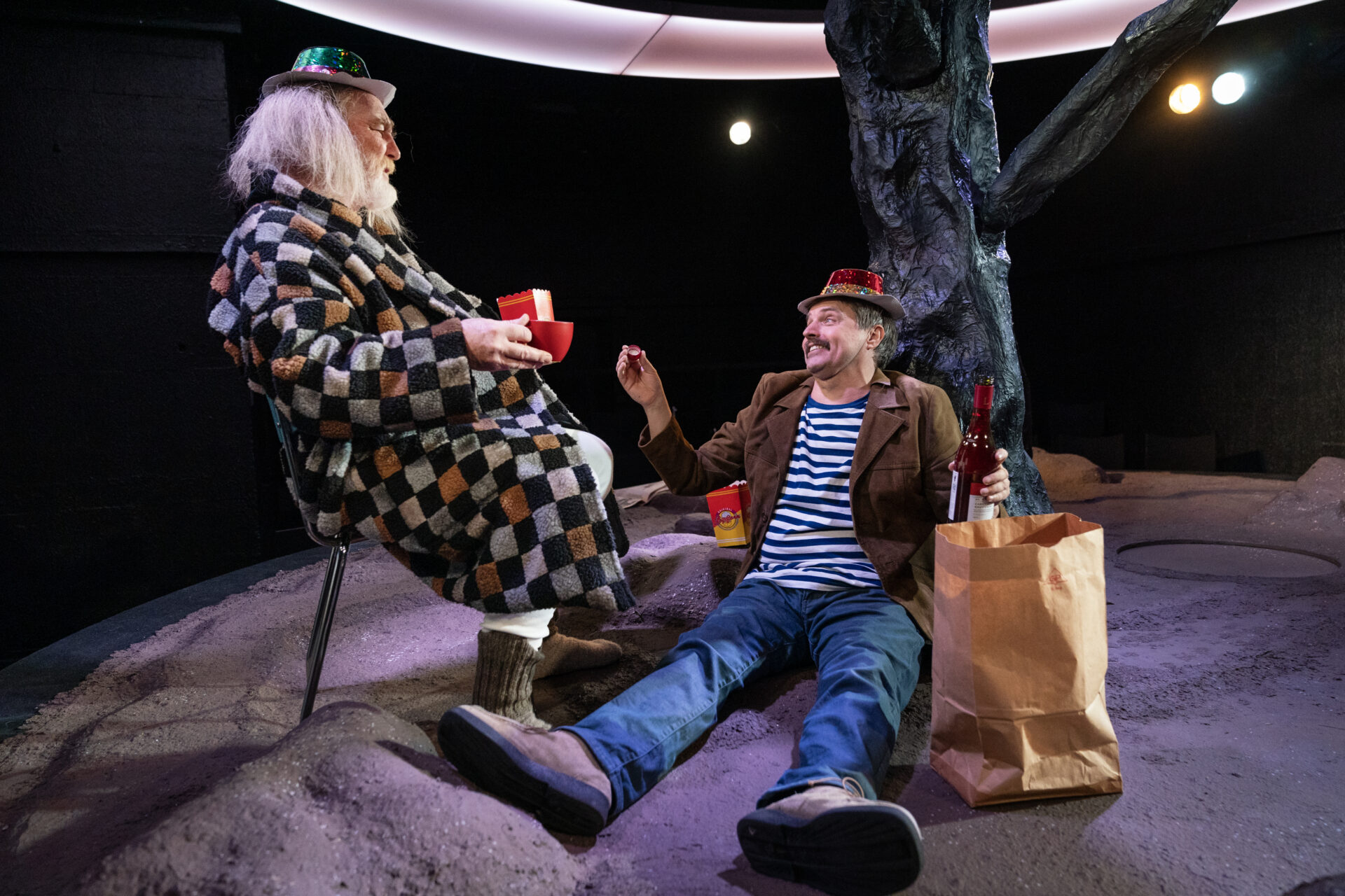 En gammel mann sitter på en stol og snakker med en yngre mann som sitter lent mot et tre.