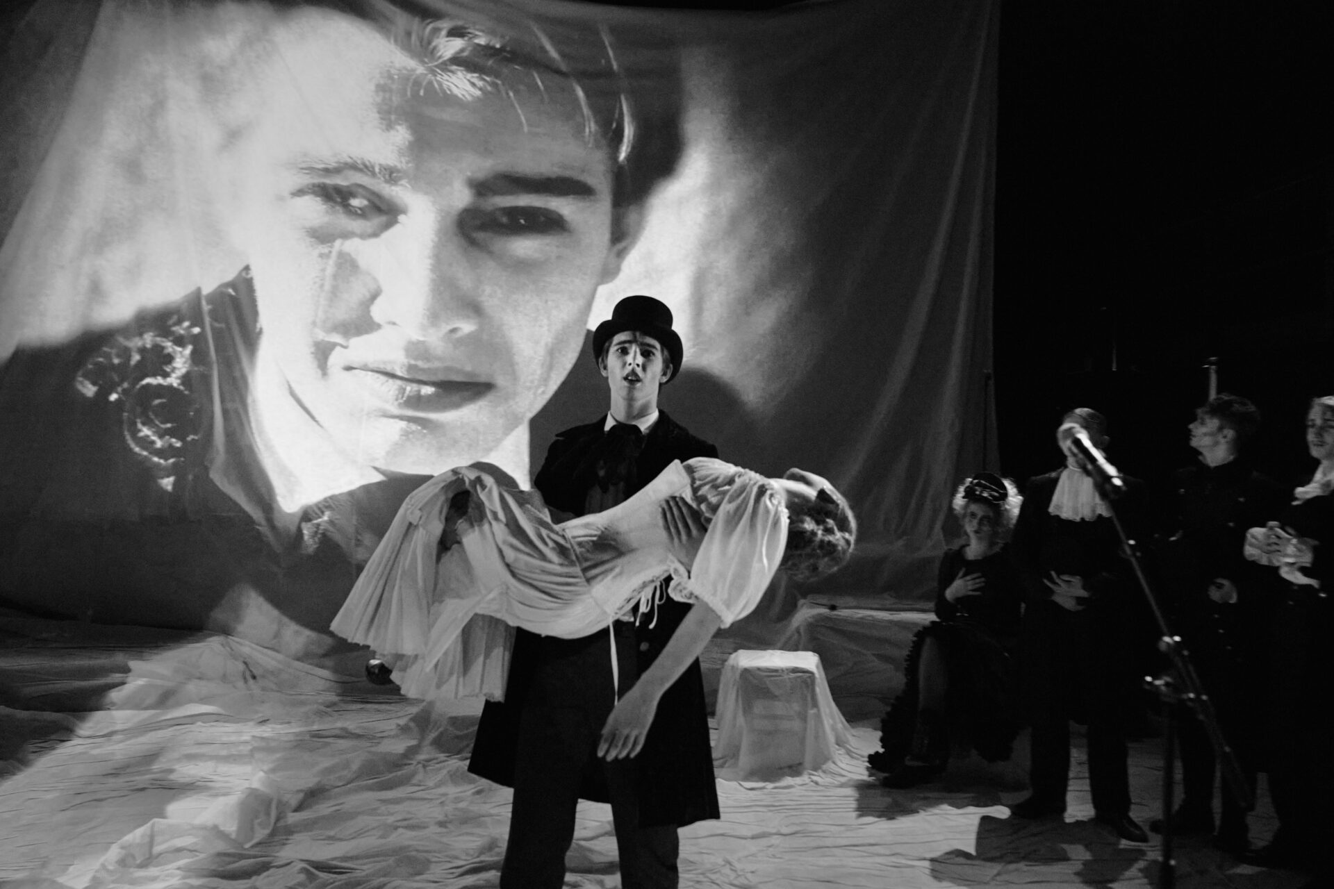 En mann bærer en død hvinne i sine armer og bak sees en gråtende mann på et filmlerret.