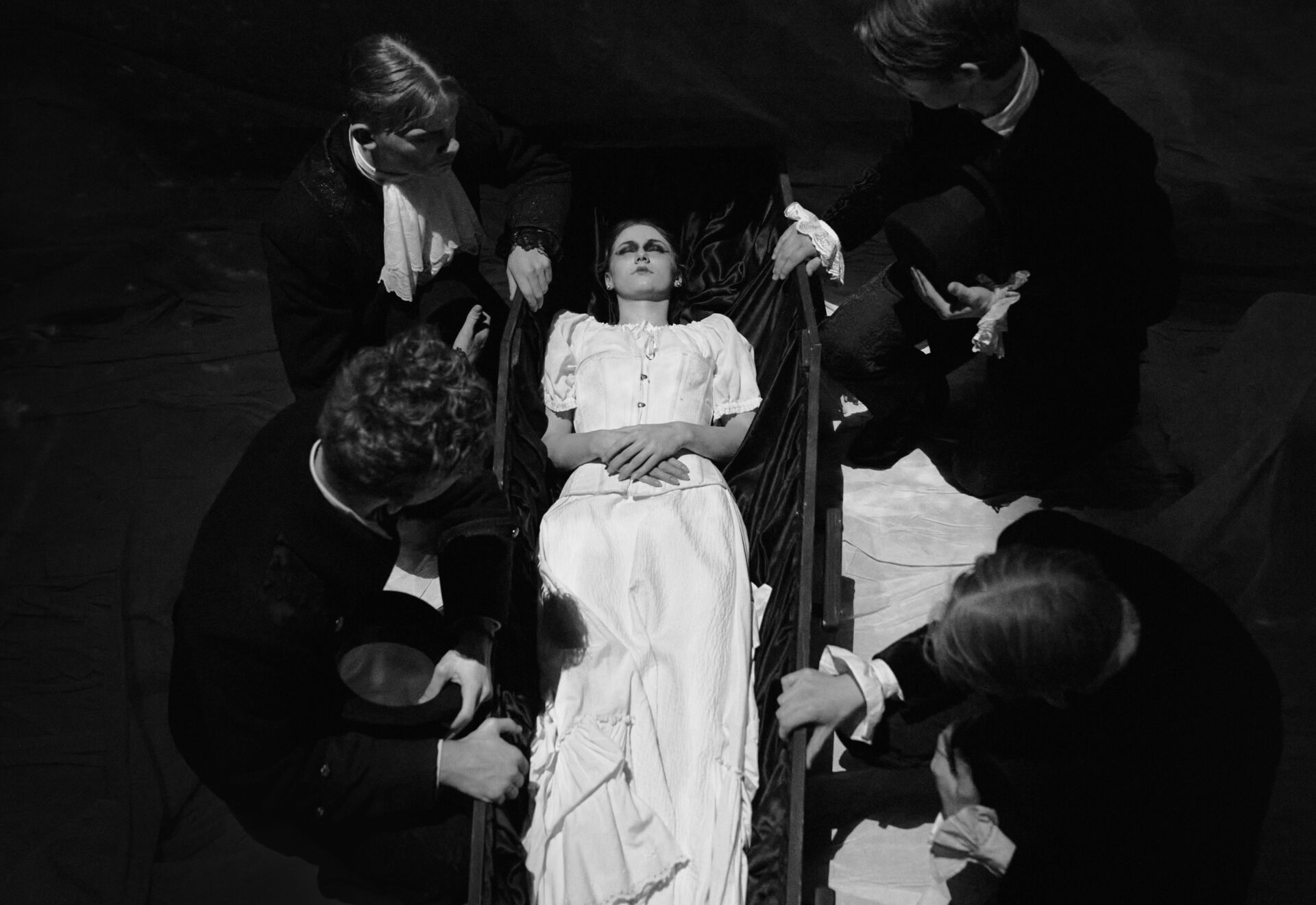 En kvinne ligger i en kiste og fire menn ser sorgfylt ned på kvinnen