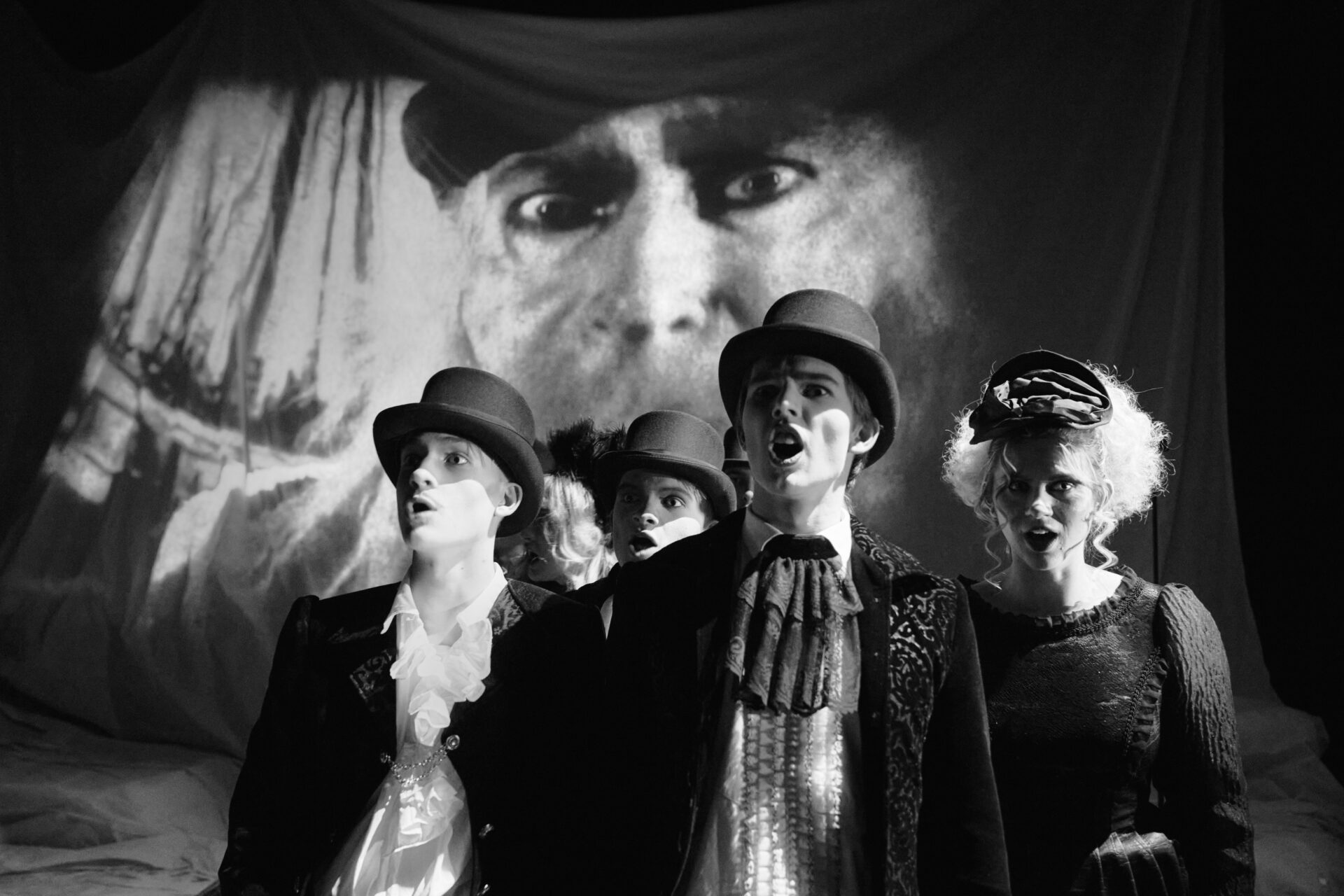 En kvinne og fire menn i klær fra 1800 tallet står foran et filmlerret med en skummel kvinne på