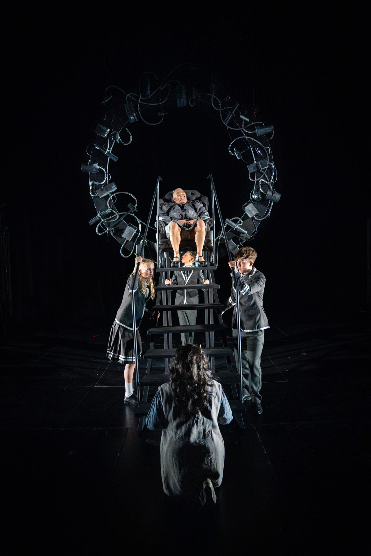 En ondskapsfull kvinne sitter på en trone laget av en lenestol mens tre ungdommer står under og holder fast i tronen. Foran kneler en kvinne