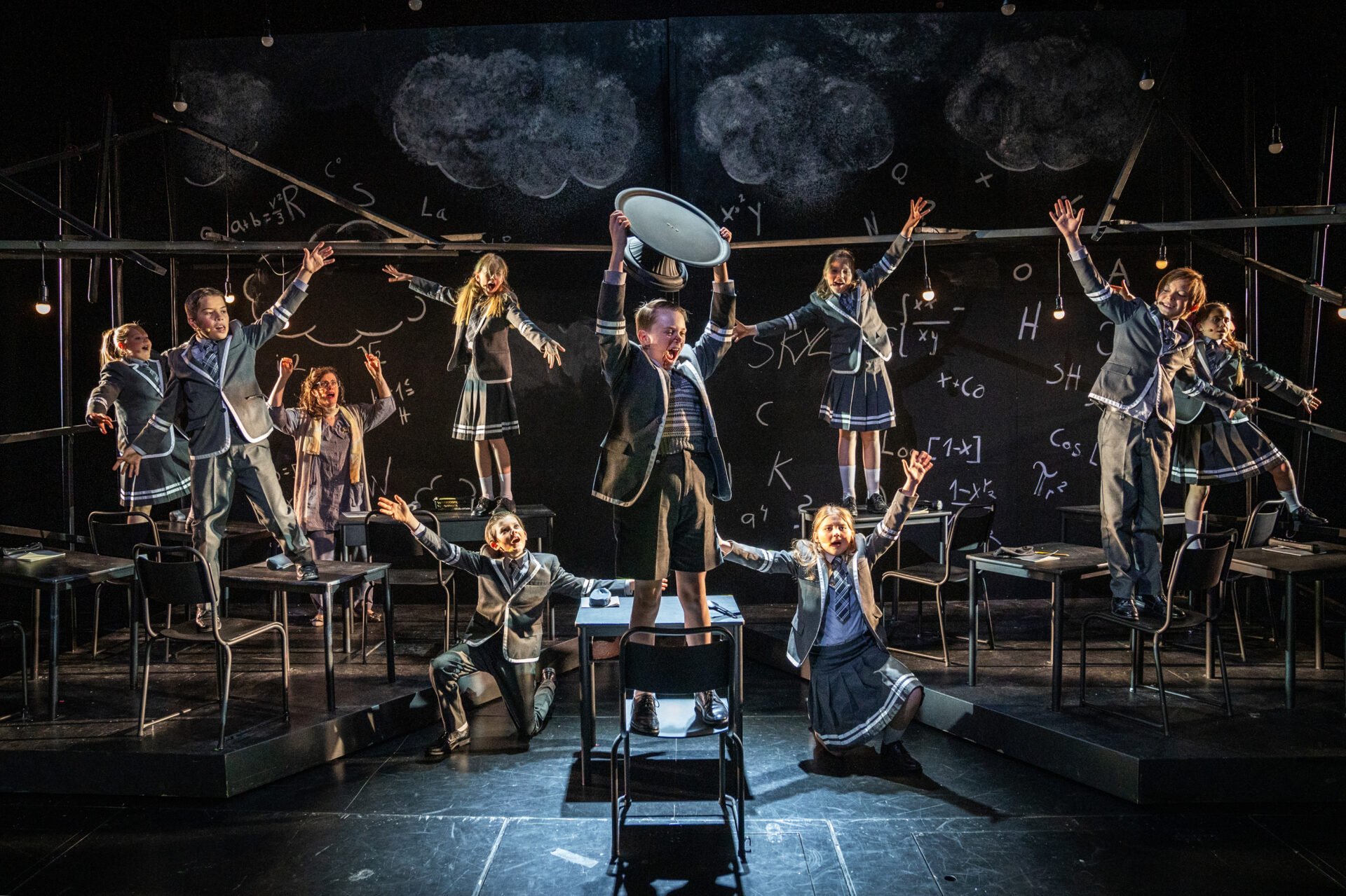 En hel skoleklasse står på pultene og heier på en gutt som holder et tomt kakefat over hodet i triumf