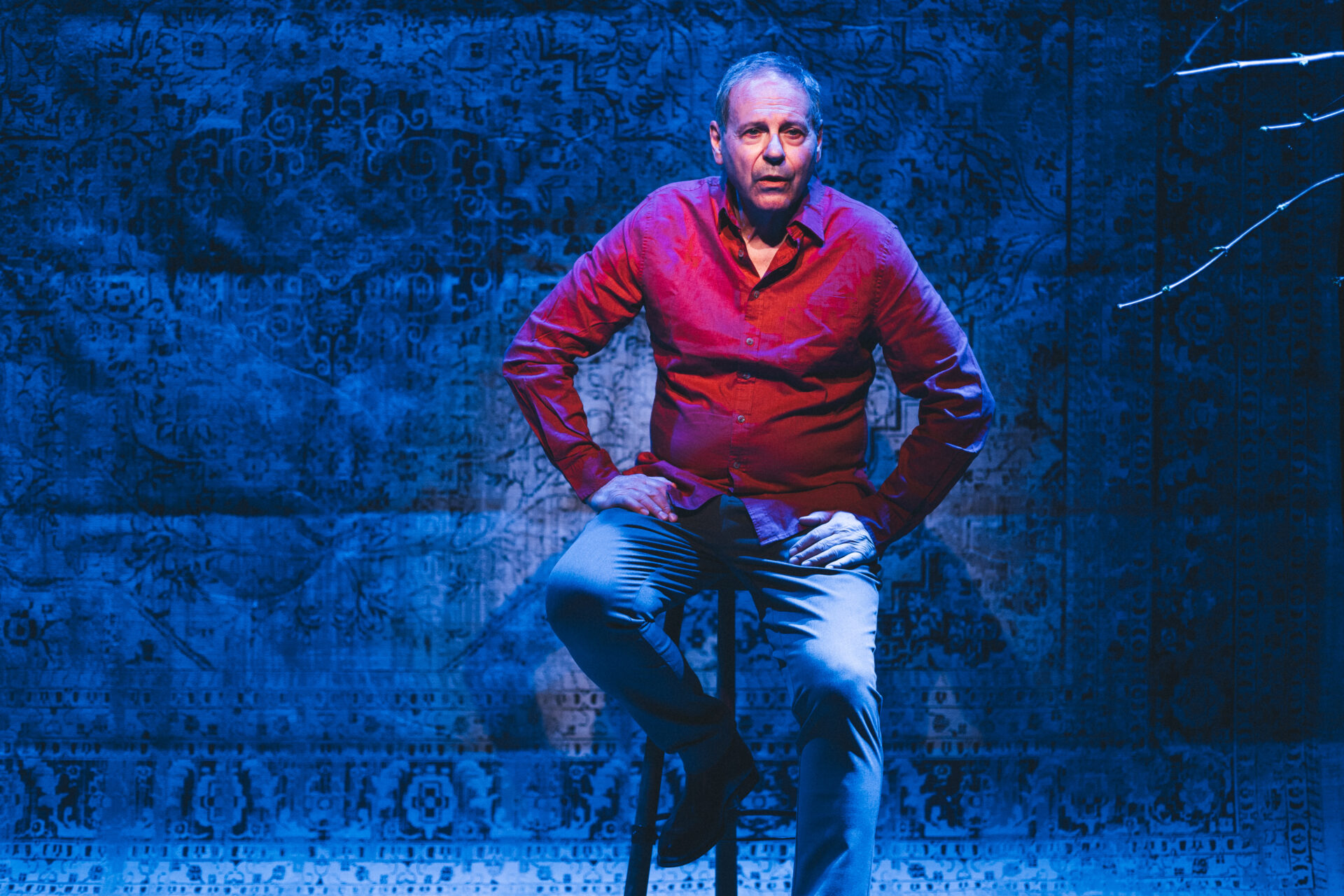 En mann i rød skjorte som sitter på en krakk på scenen, og titter utover