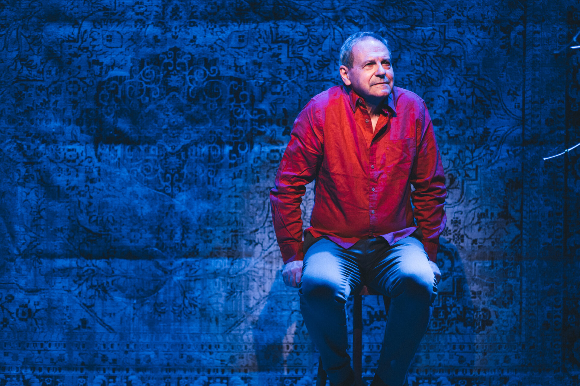 En mann i rød skjorte som sitter på en krakk på scenen, og titter utover