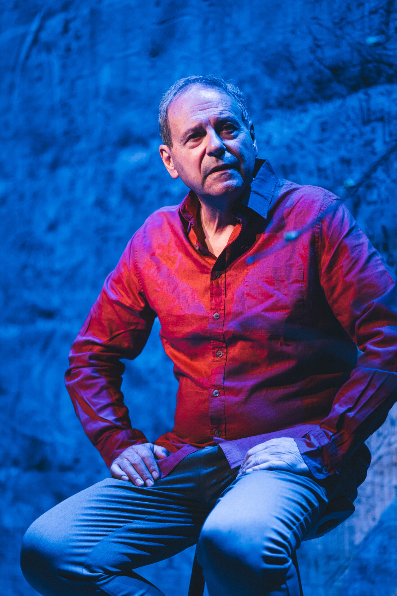 En mann i rød skjorte som sitter på en krakk på scenen, og titter utover