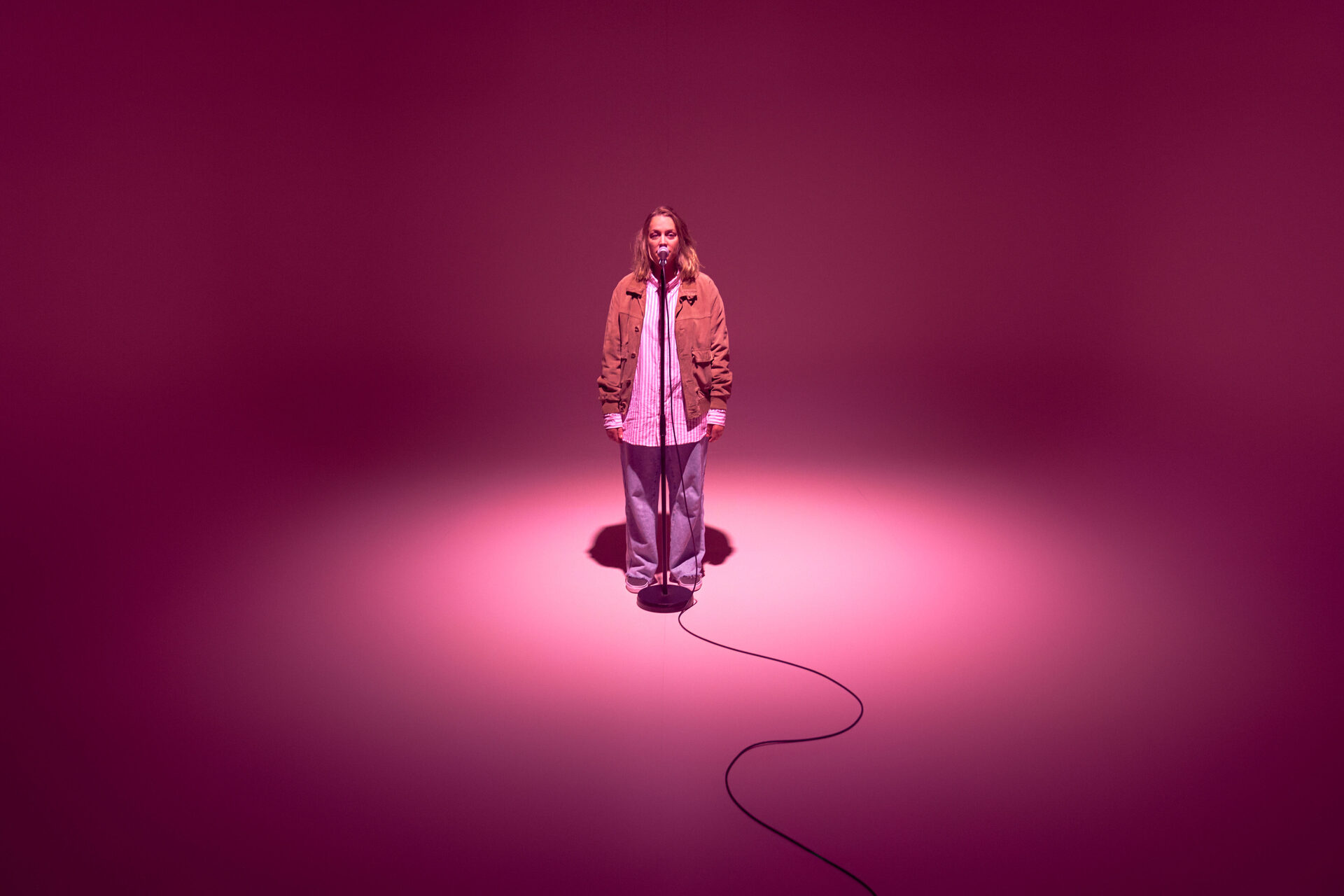 En kvinne står alene på en rosa scene foran en mikrofon.