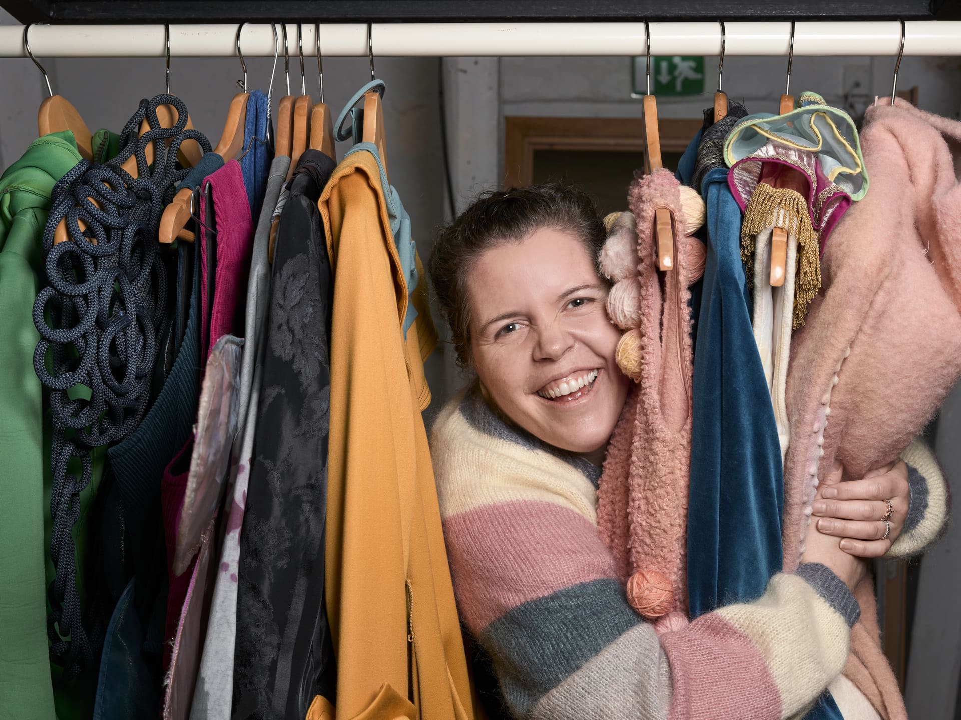 En smilende dame holder rundt fargerike kostymer som henger på et klesstativ.