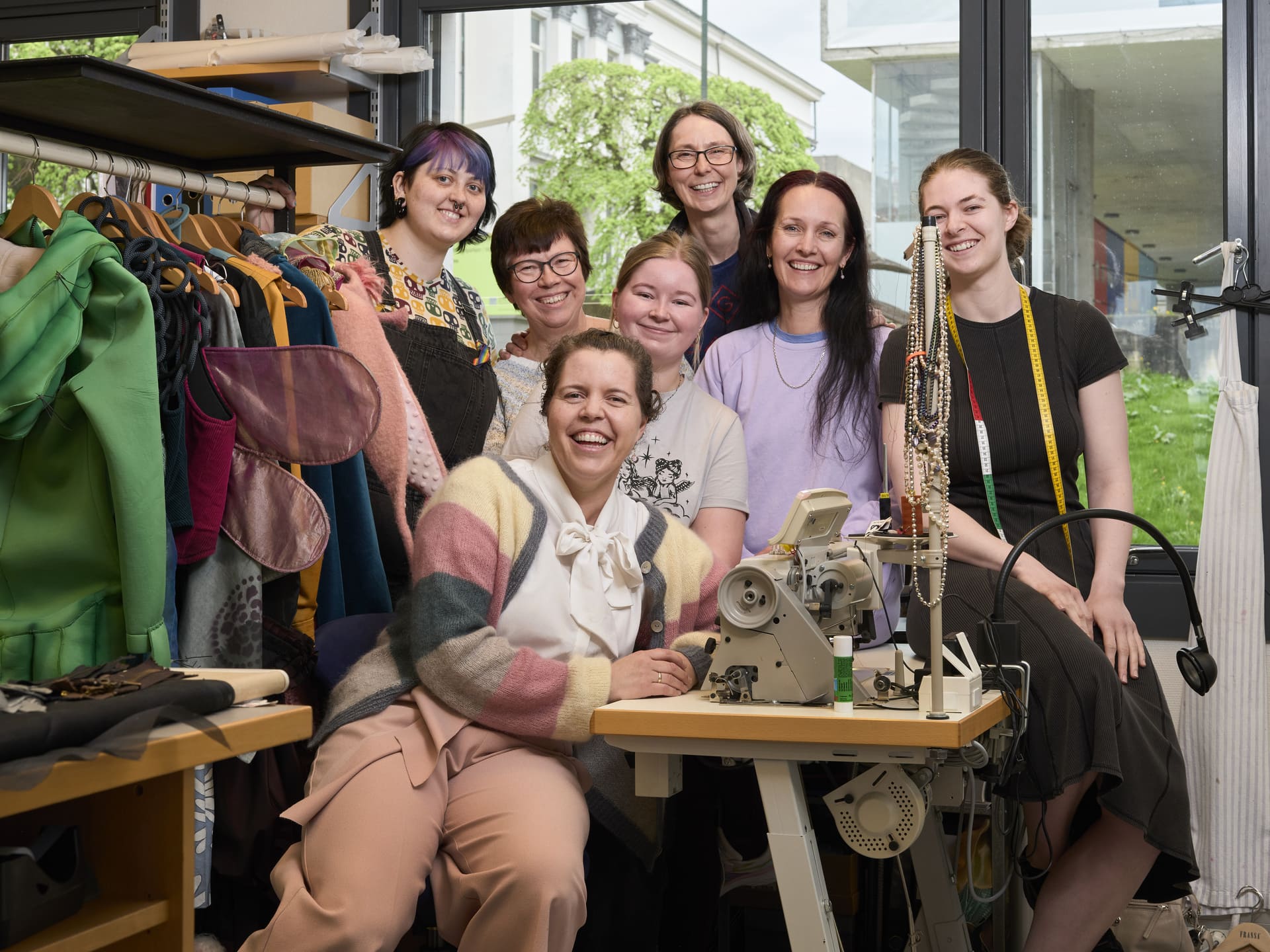 Syv kvinner står og sitter blant kostymer, symaskin og verktøy.