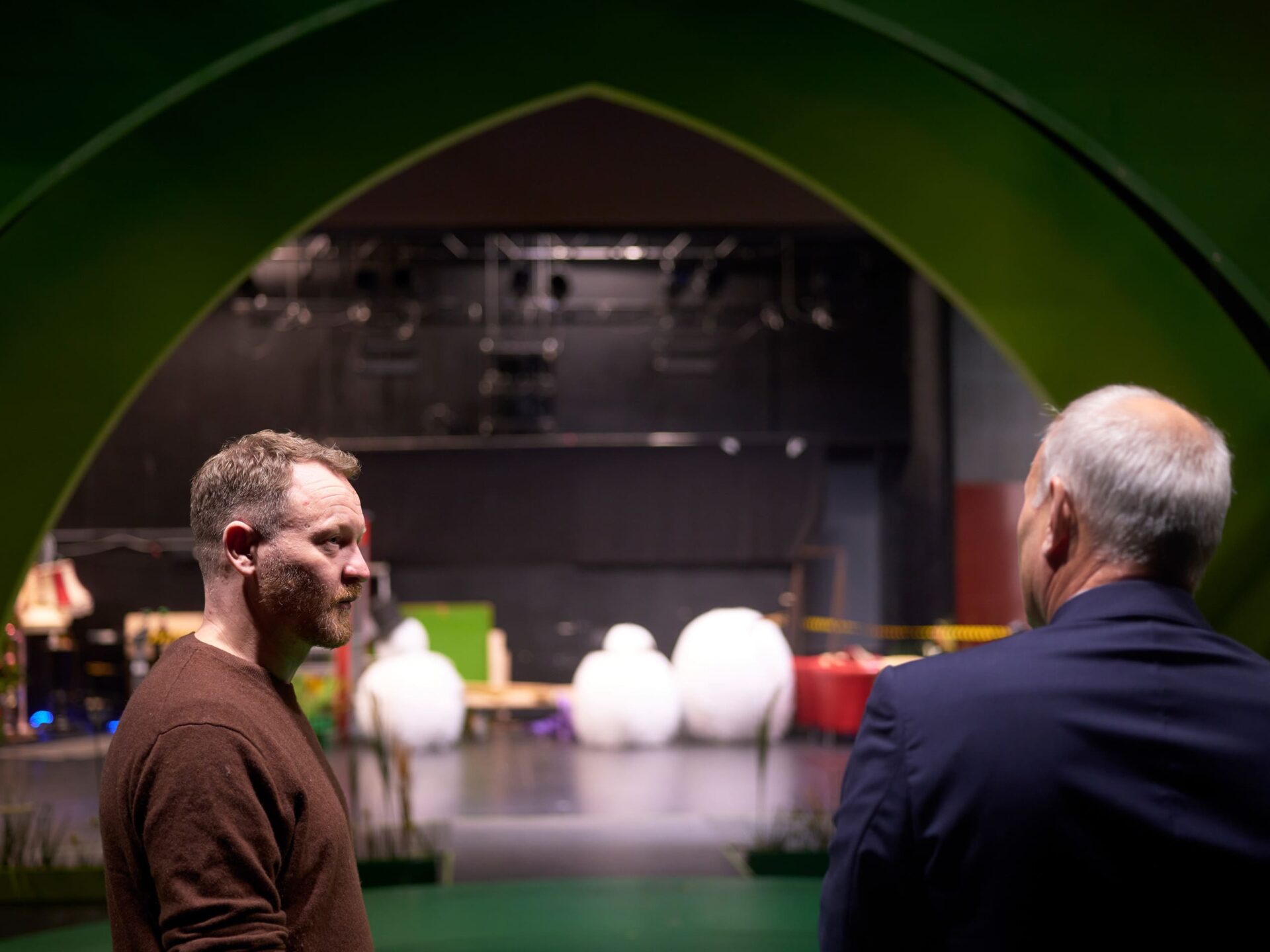 To menn prater sammen. I bakgrunnen er en teaterscene.