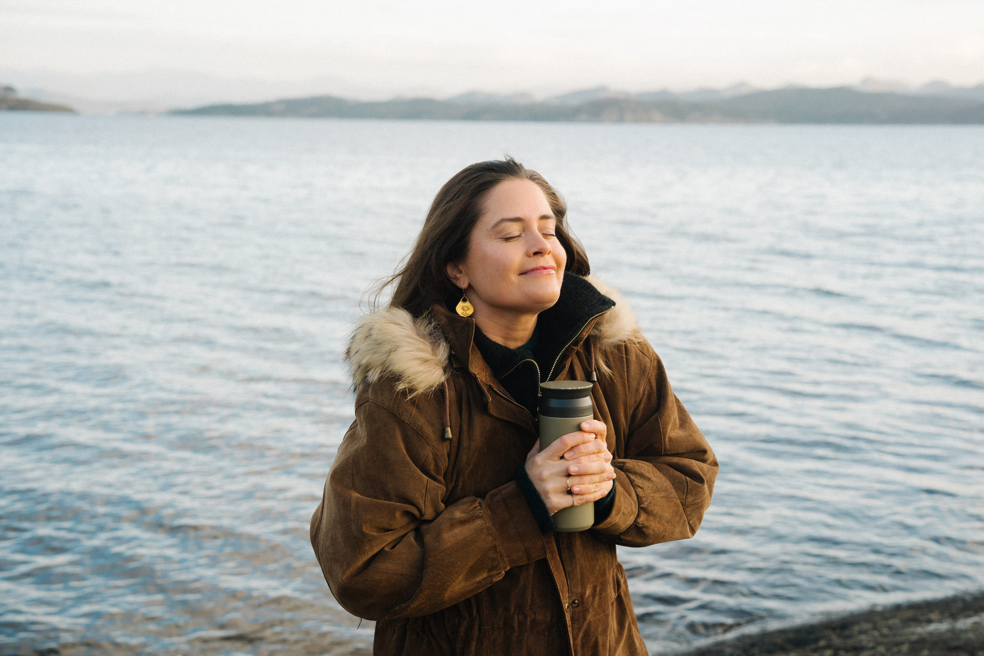 Kvinne står med kaffekopp ved havet.