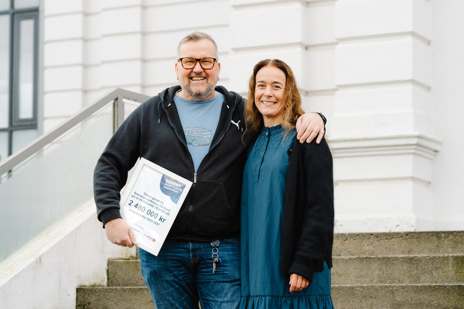 En smilende mann har armen rundt en smilende kvinne. Mannen holder en innrammet bekreftelse på pengegave.