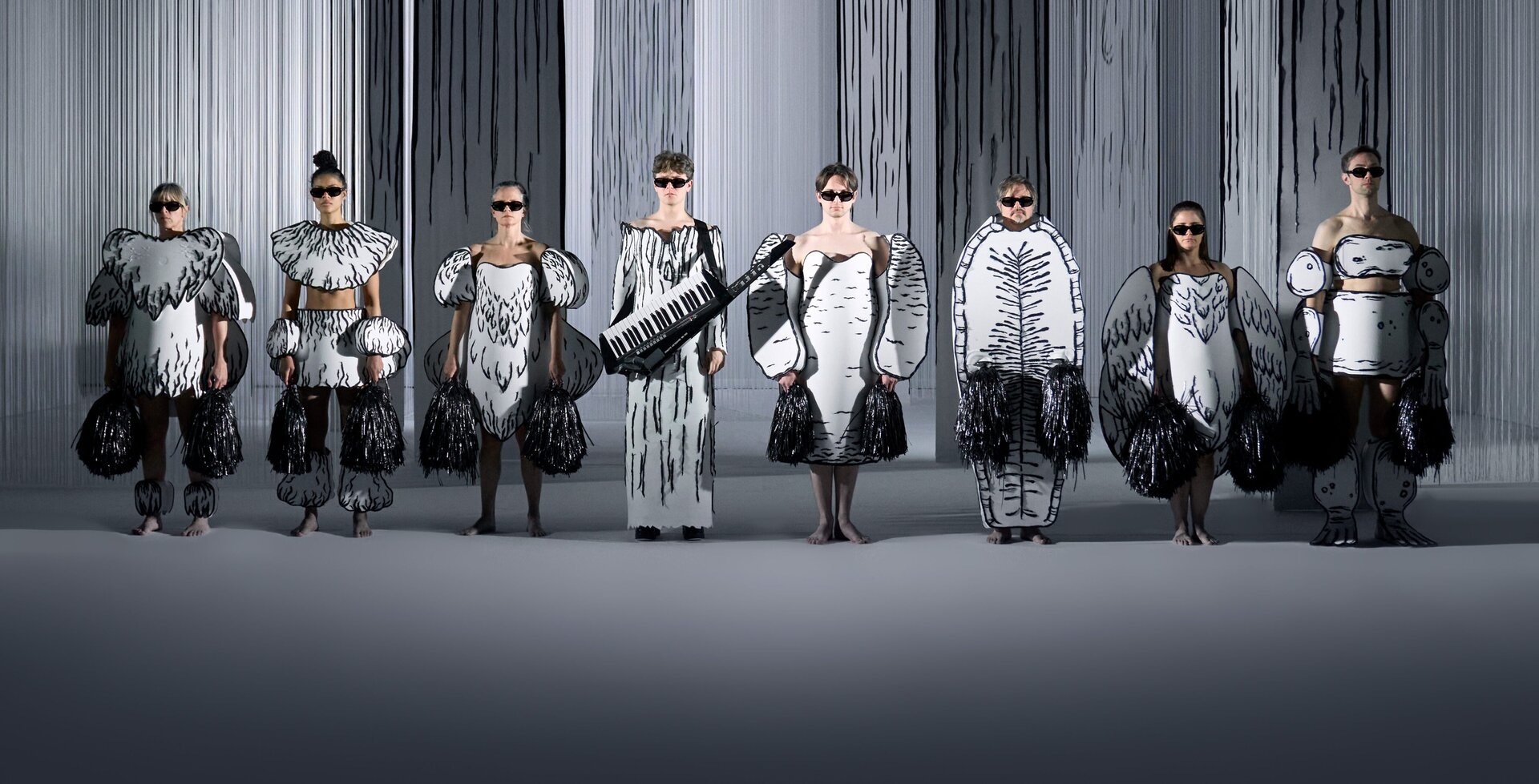 Syv skuespillere utkledd som sort-hvitt kulltegnede dyr uten masker.