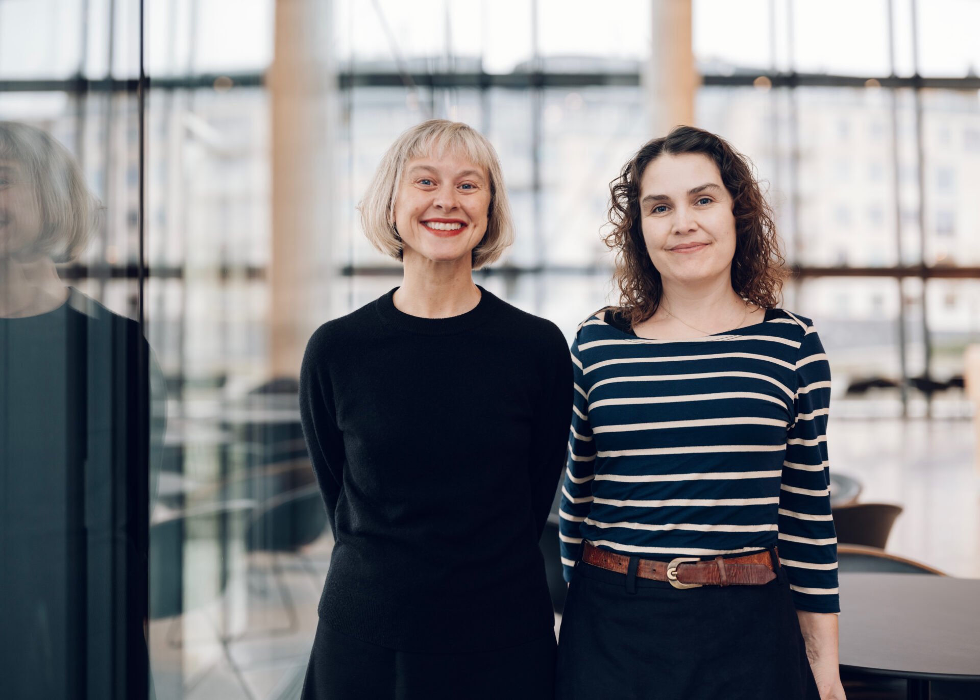 To smilende kvinner står foran en glassvegg.