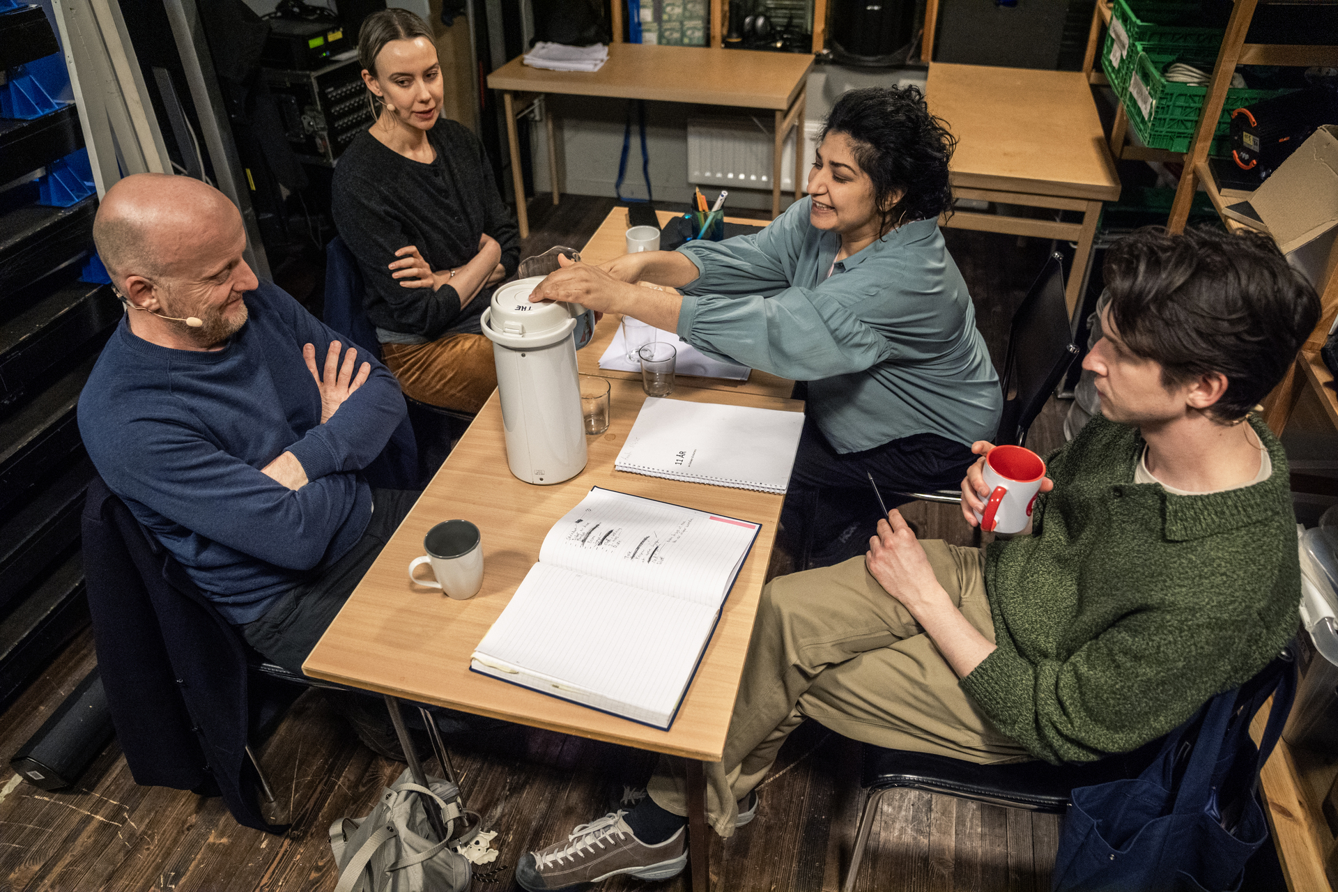 Fire skuespillere sitter rundt et lite bord omgitt av hyller, skap og teknisk utstyr.